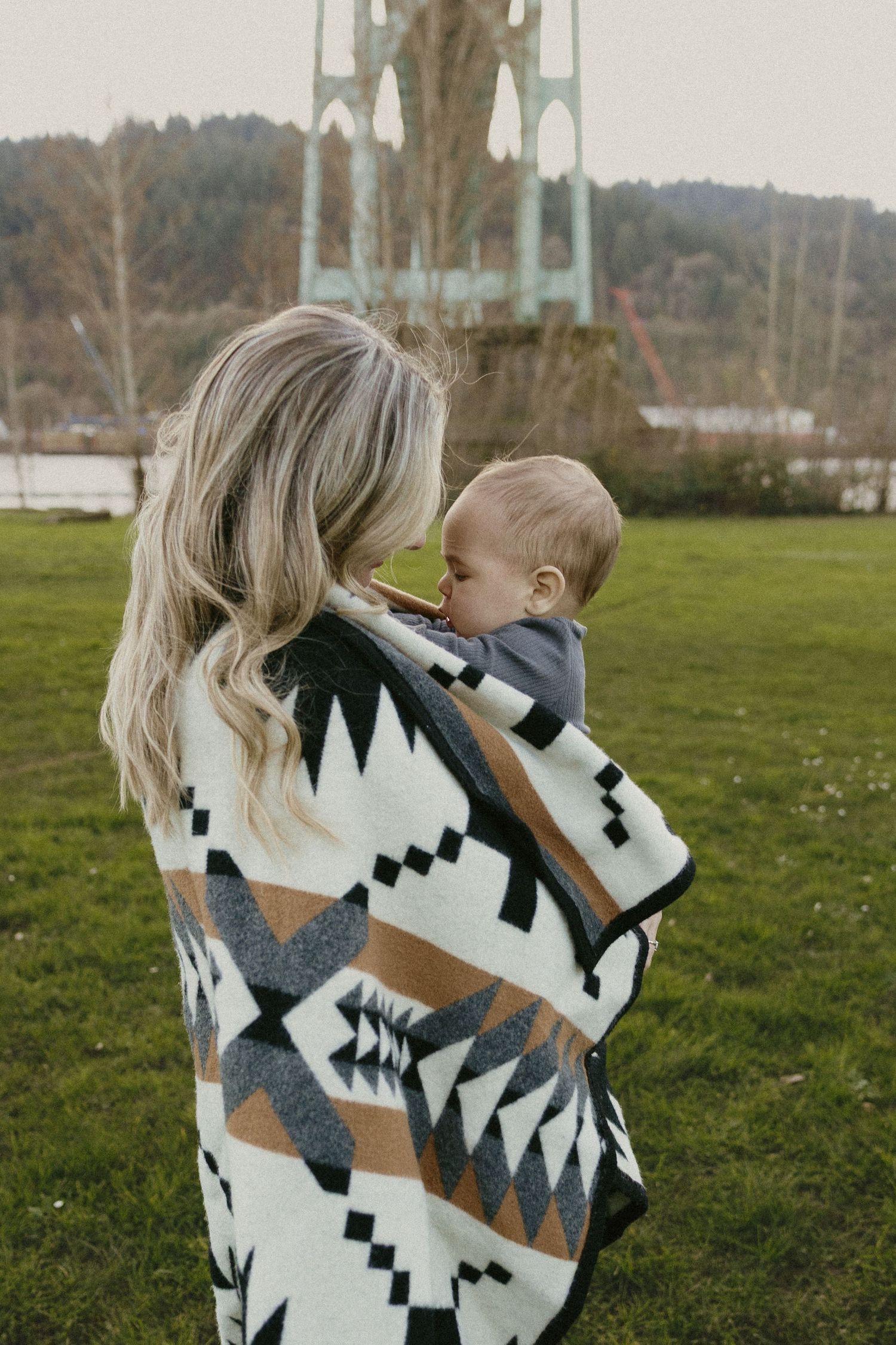 Mother wrapped in geometric patterned blanket holding baby while standing in park with bridge in background.