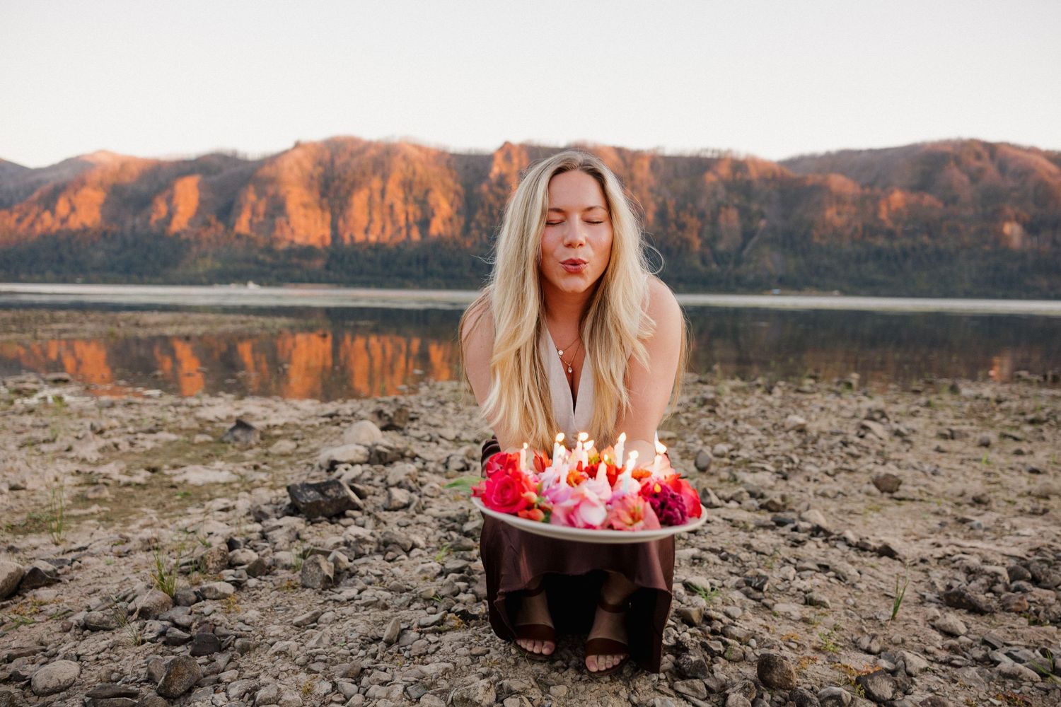 Blonde woman blowing out candles on a floral cake by a mountain lake at sunset.