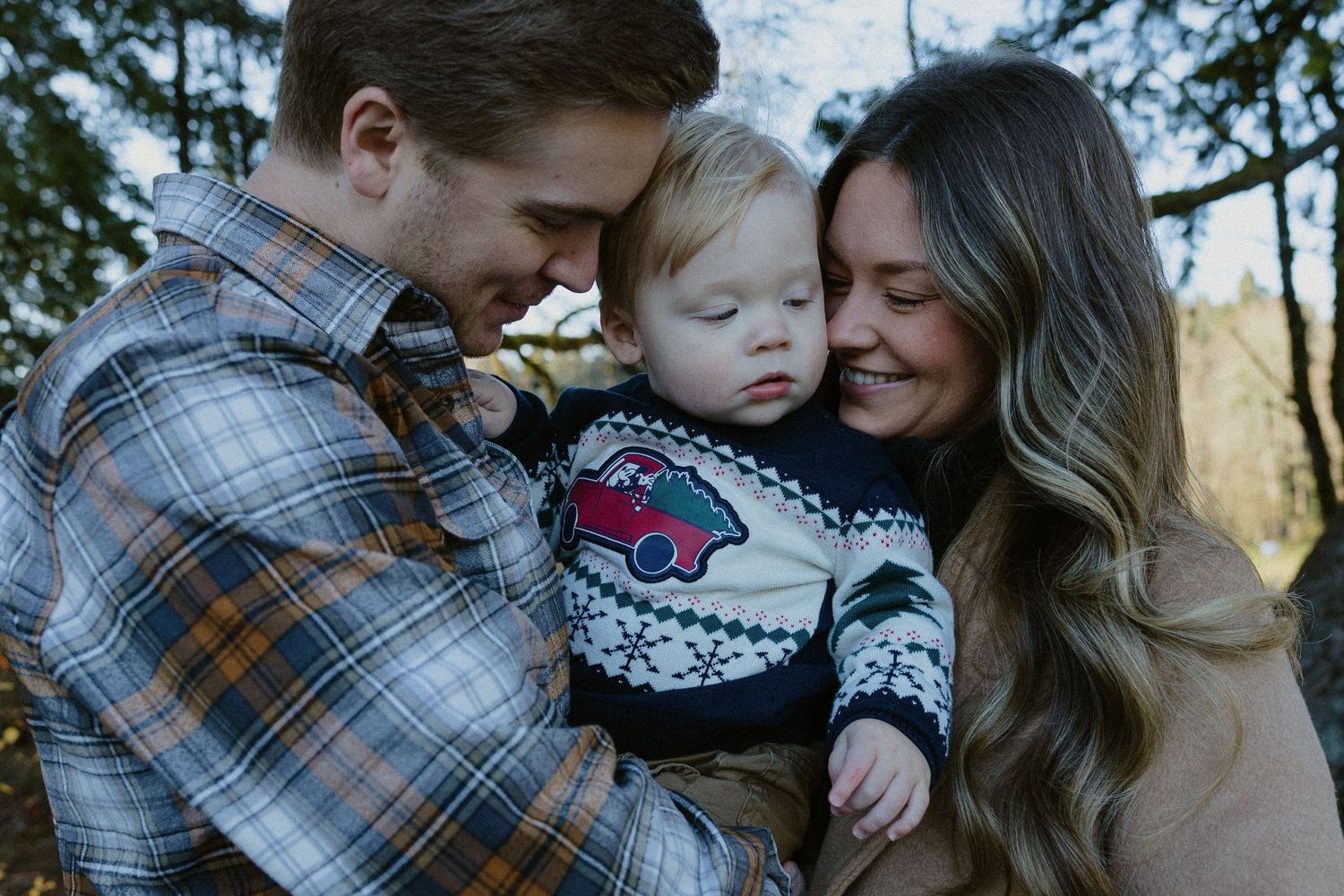 Young couple sharing tender moment with toddler in holiday sweater among autumn forest trees.