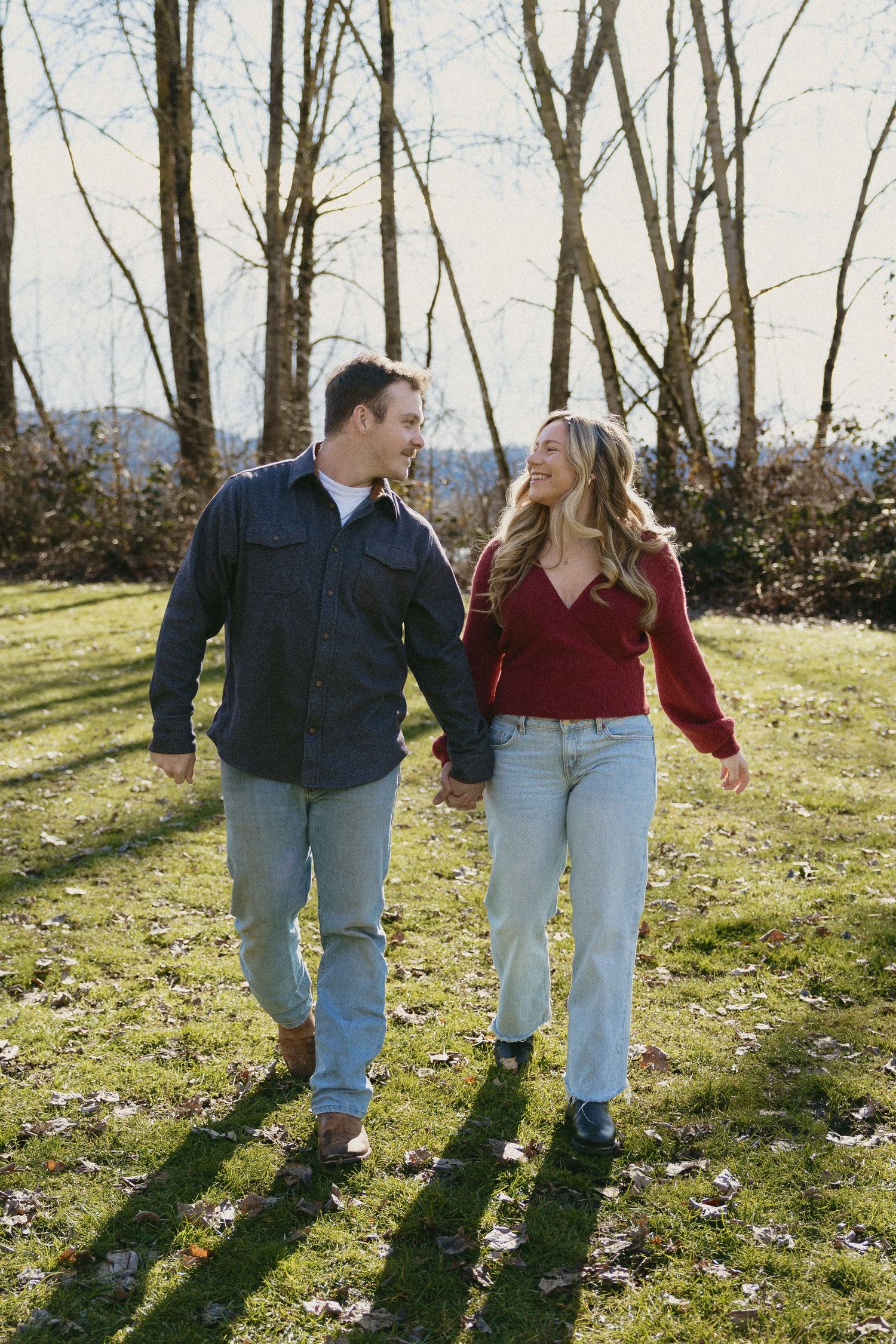 Couple walking hand in hand on green lawn smiling at each other with bare winter trees.