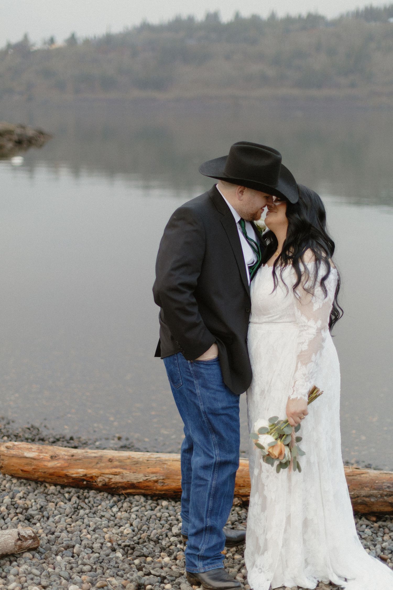 Bride and groom kissing on a rocky shoreline, groom in cowboy hat, bride holding a bouquet.