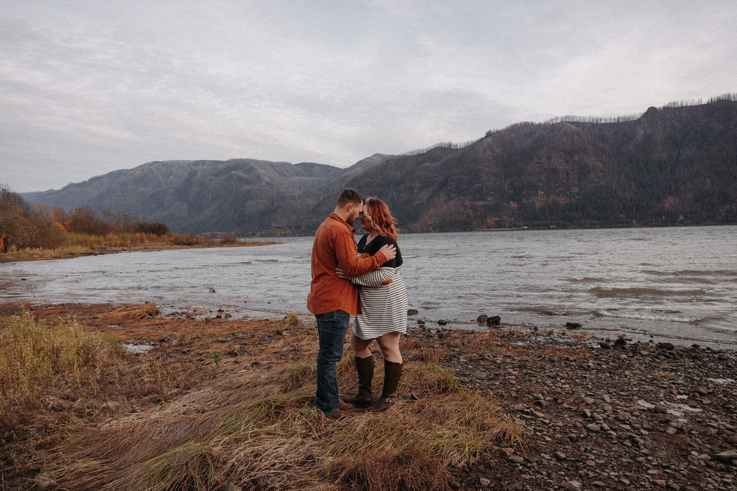 Couple hugging tenderly on a rocky riverside with mountains in the background.