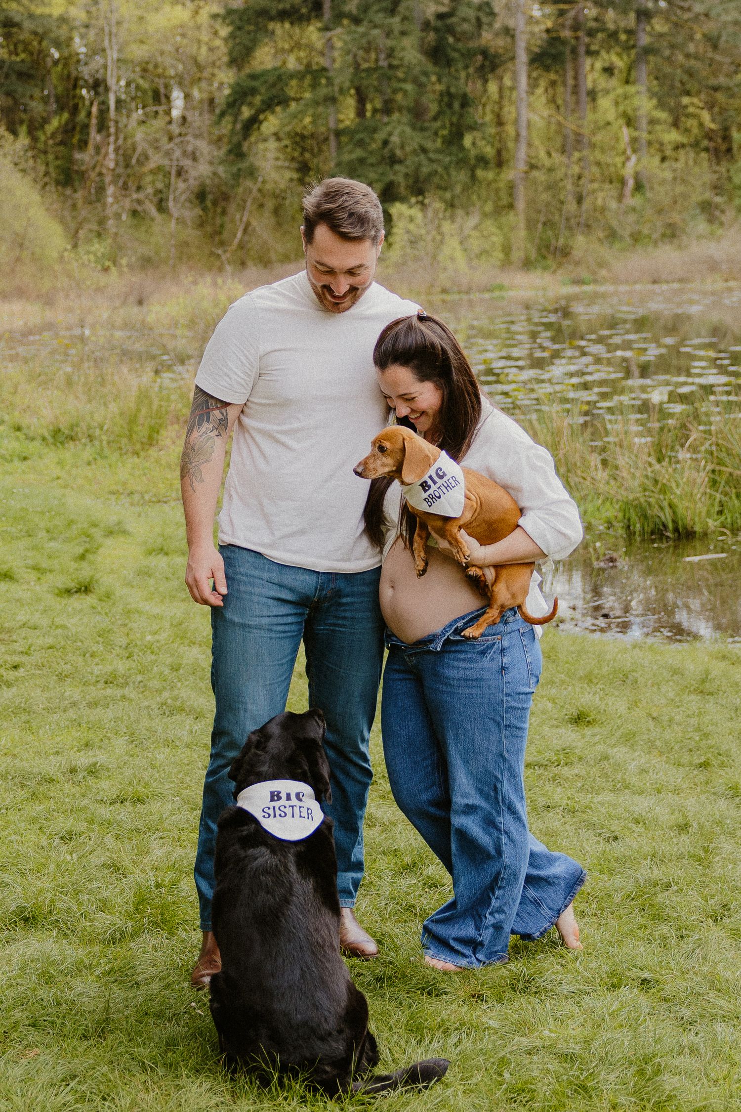 Couple with two dogs wearing bandanas by a pond outdoors.