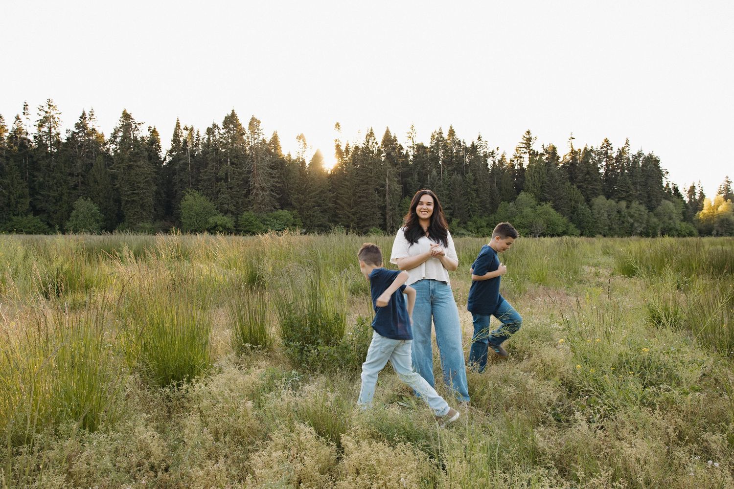 A smiling mother walks through a golden-hour meadow with her two boys running beside her near a forest.