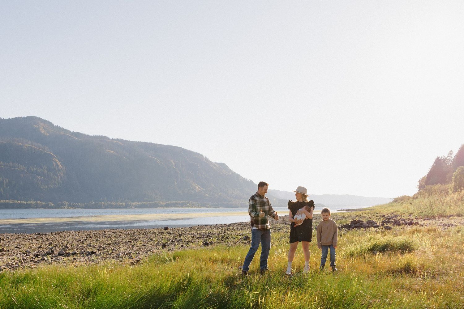 A family of four stands together on a grassy riverbank with scenic mountain views in the background.