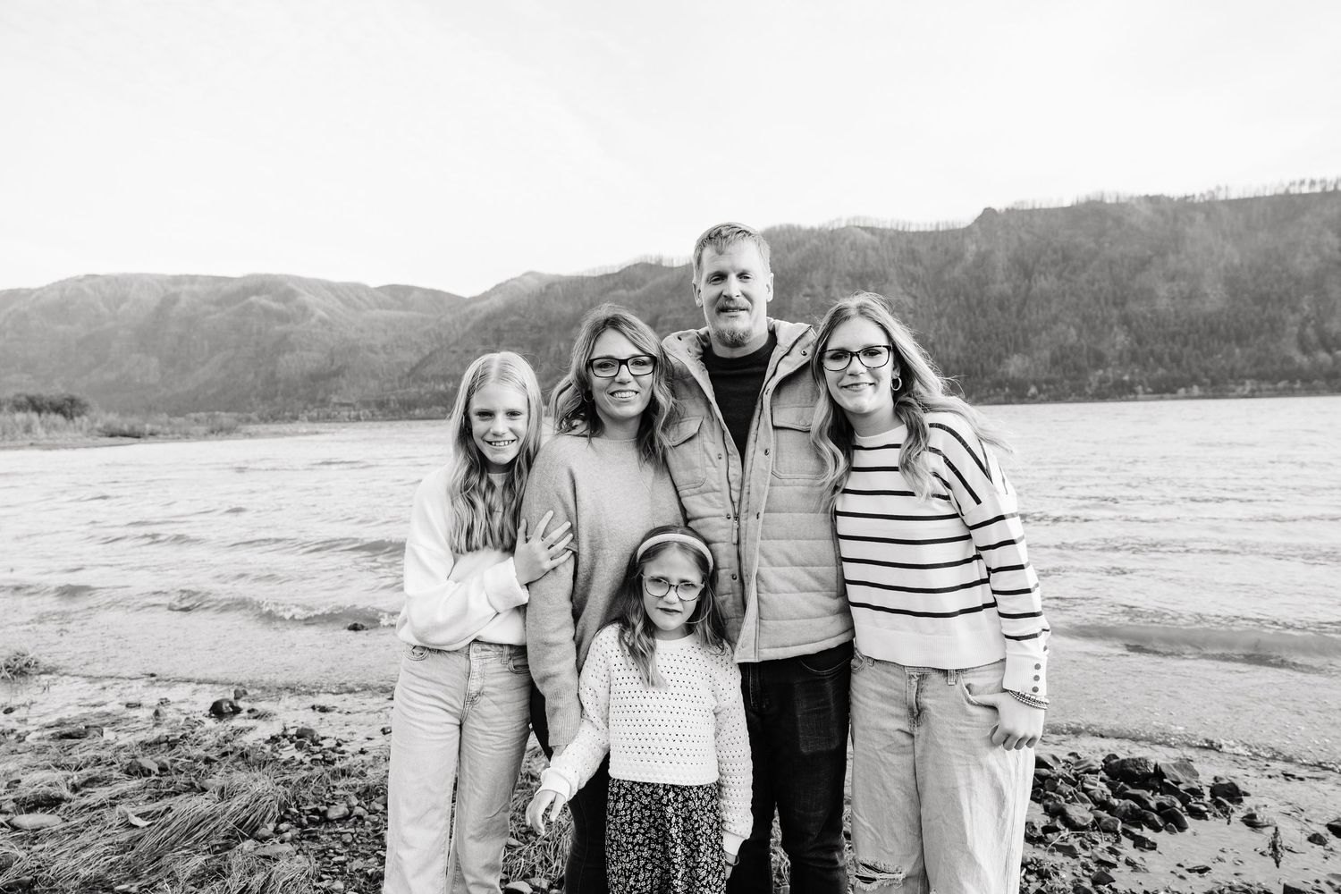 Black and white family portrait of five on a rocky riverbank with mountain backdrop.