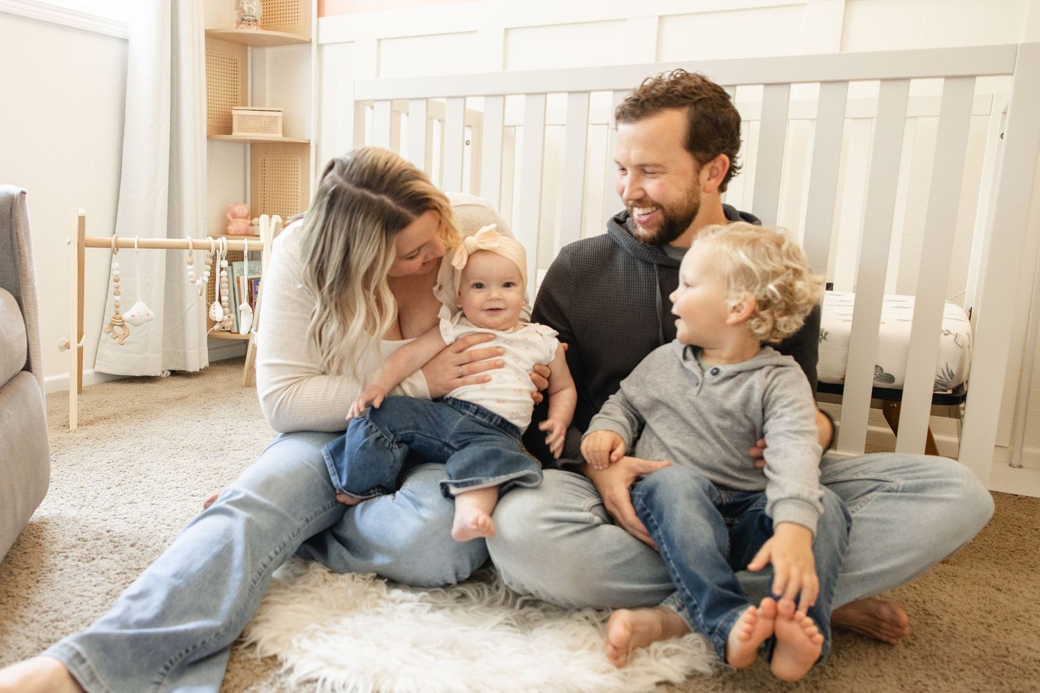 Young family sitting together on nursery floor near white crib with two toddlers.