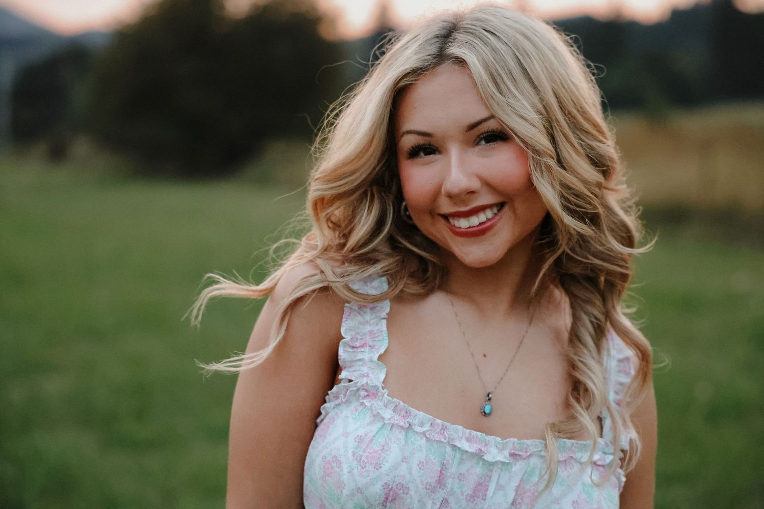 Smiling blonde woman in floral dress posing outdoors with green fields and mountains behind her.