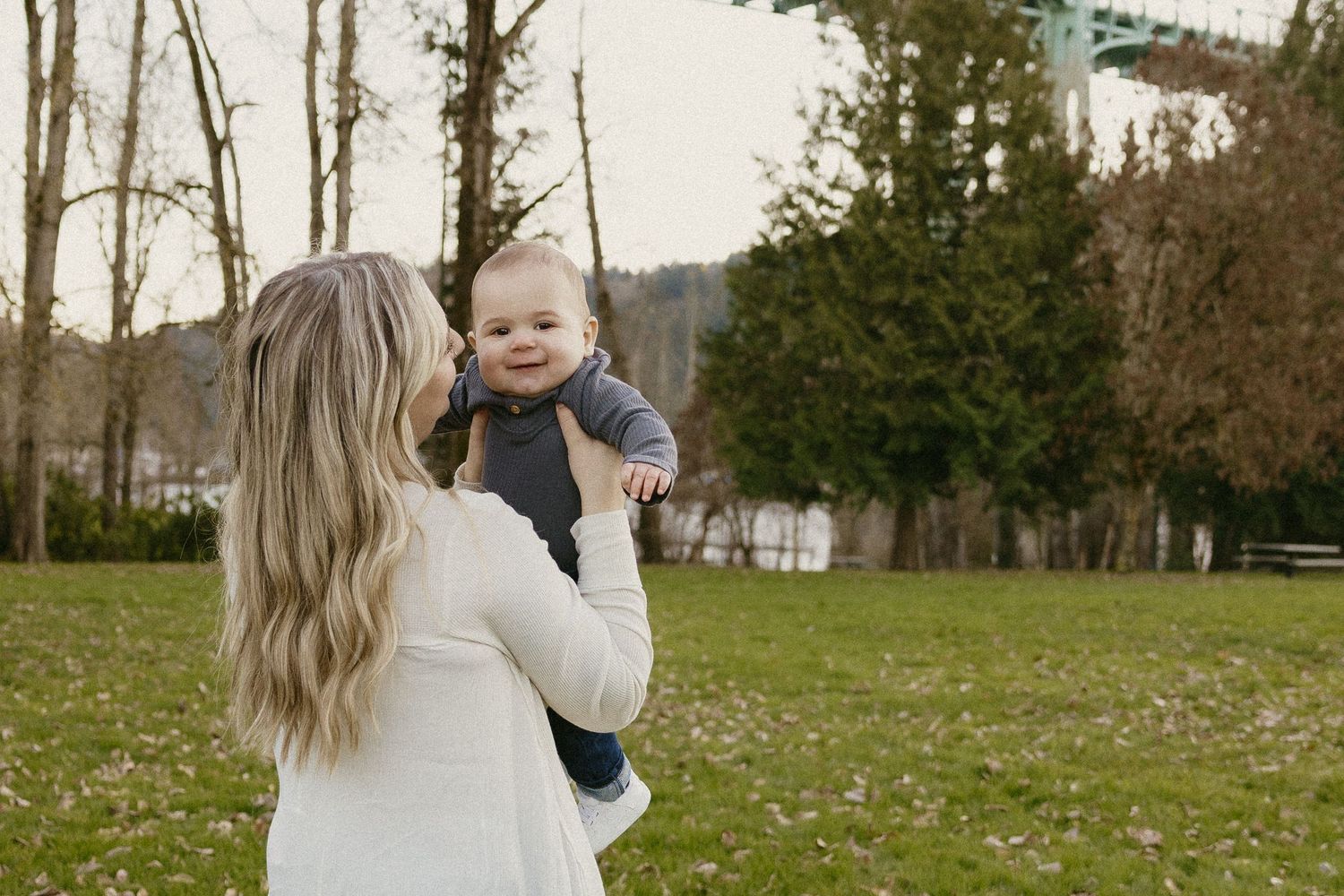 Blonde mother lifting smiling baby boy in the air at Cathedral Park near St. Johns Bridge Portland.