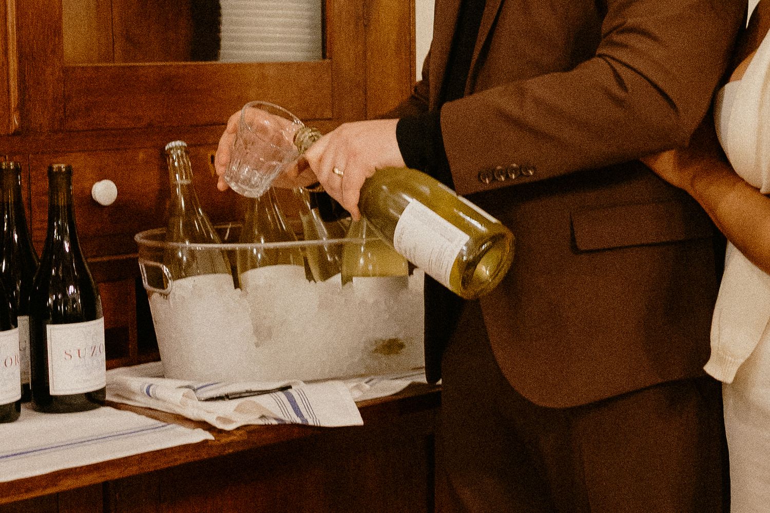 Groom in brown suit pouring white wine into ice bucket at wedding reception.