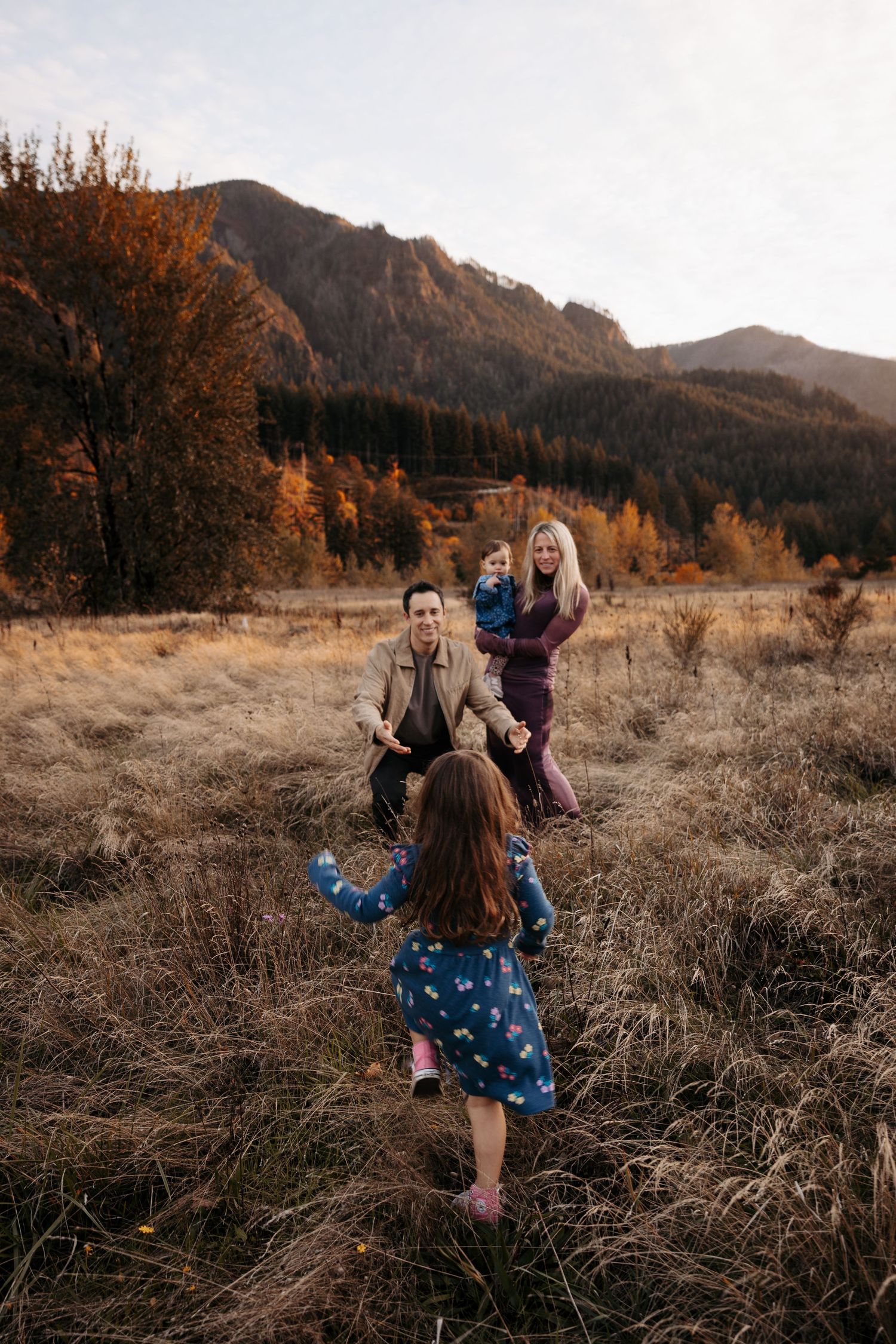 Family walking in golden autumn meadow with mountain backdrop, young girl running toward parents.