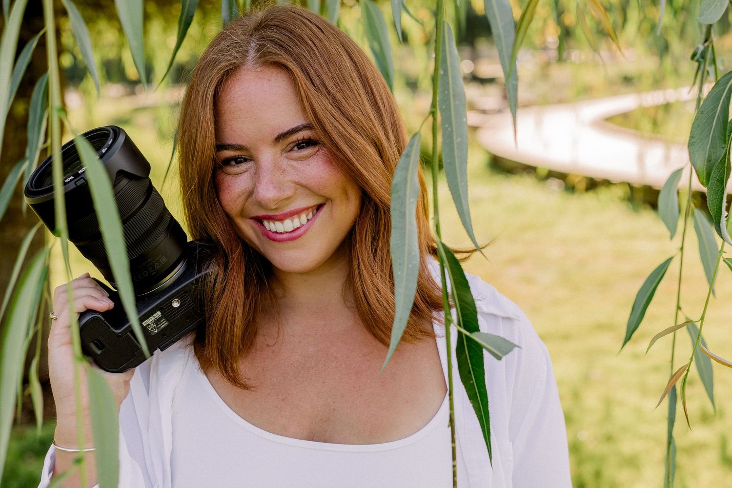 Smiling redhead woman holding a camera among willow tree branches in a sunny outdoor park setting.