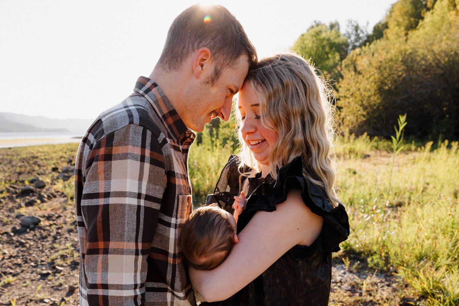 A couple leans foreheads together lovingly while holding their newborn baby in a sunny outdoor setting.
