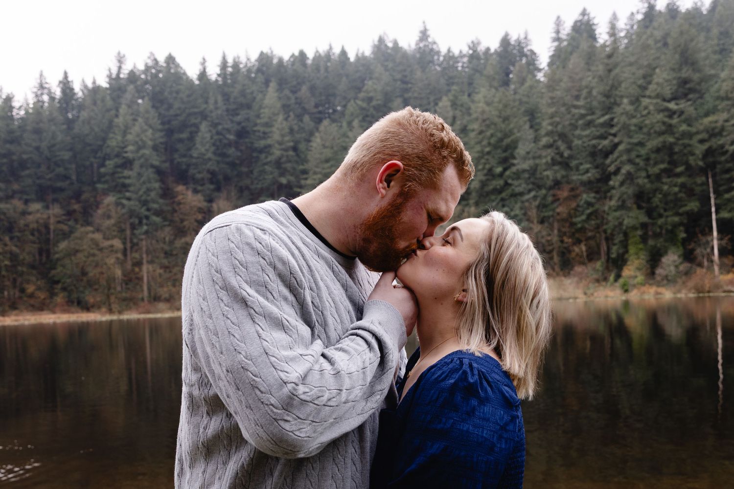Couple sharing a tender kiss by a misty forest lake.