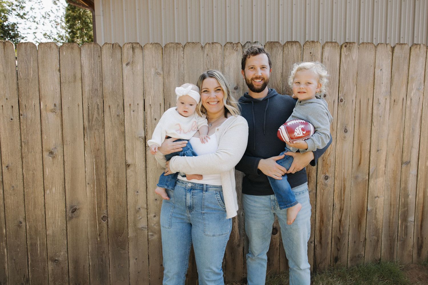 Young family of four smiling outdoors by wooden fence, parents each holding a baby and toddler.