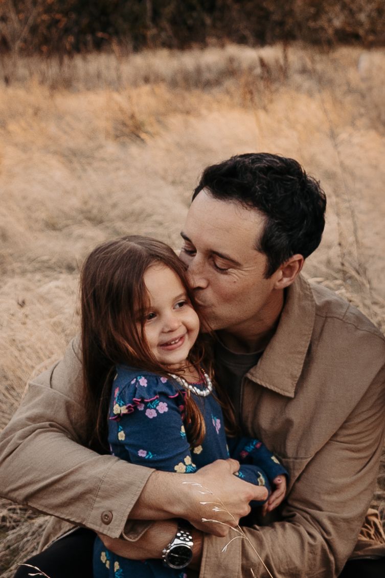 A dark-haired father kisses his smiling young daughter in a dry autumn grass field.