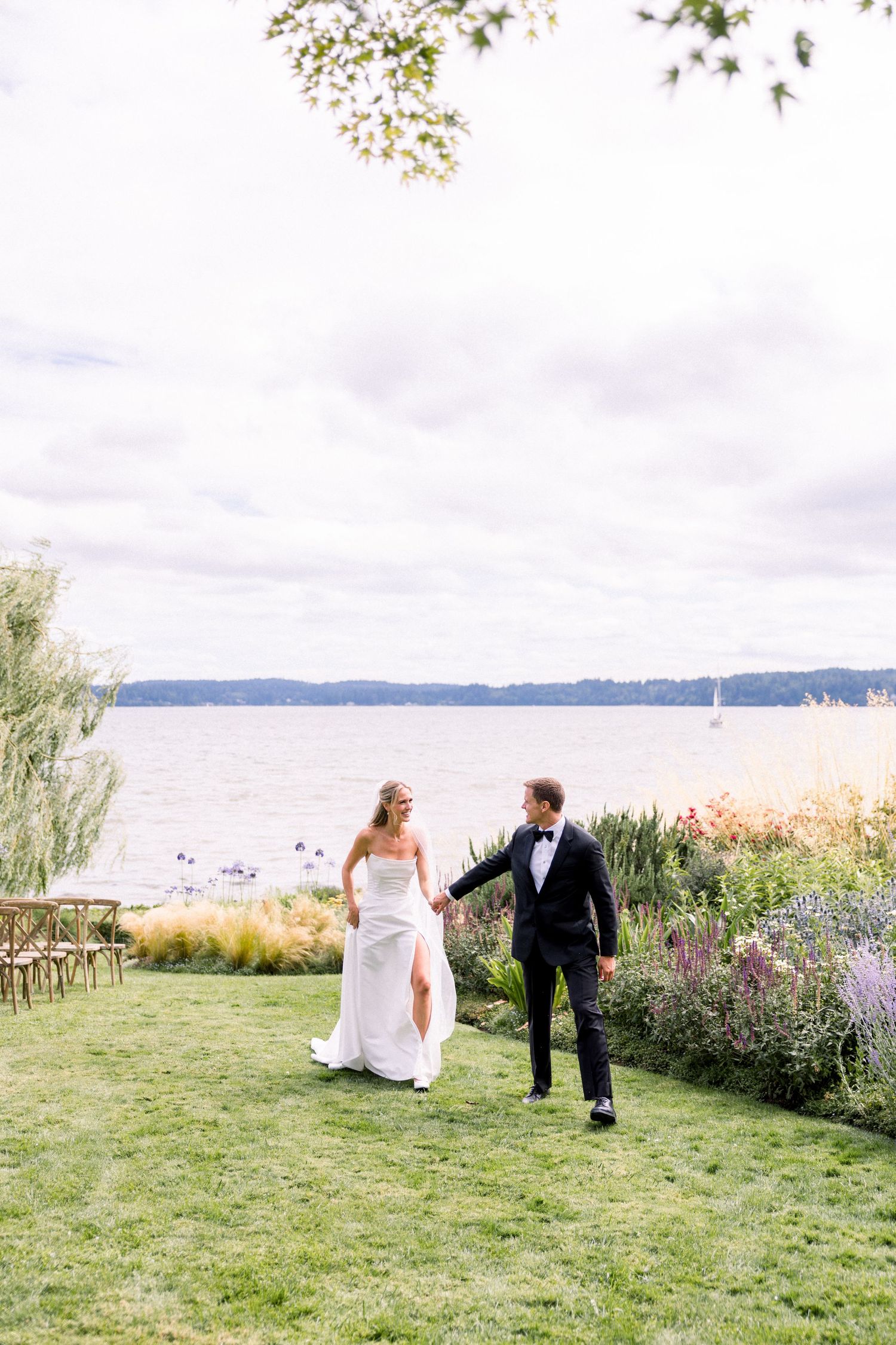 Bride and groom hold hands walking on lush lawn by waterfront garden, bride in white gown, groom in black tuxedo.