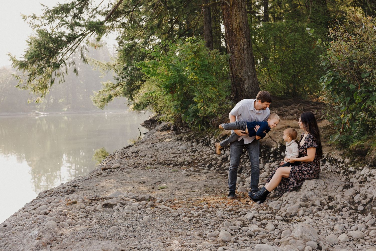 Family exploring rocky riverbank near tall pine trees on misty day.