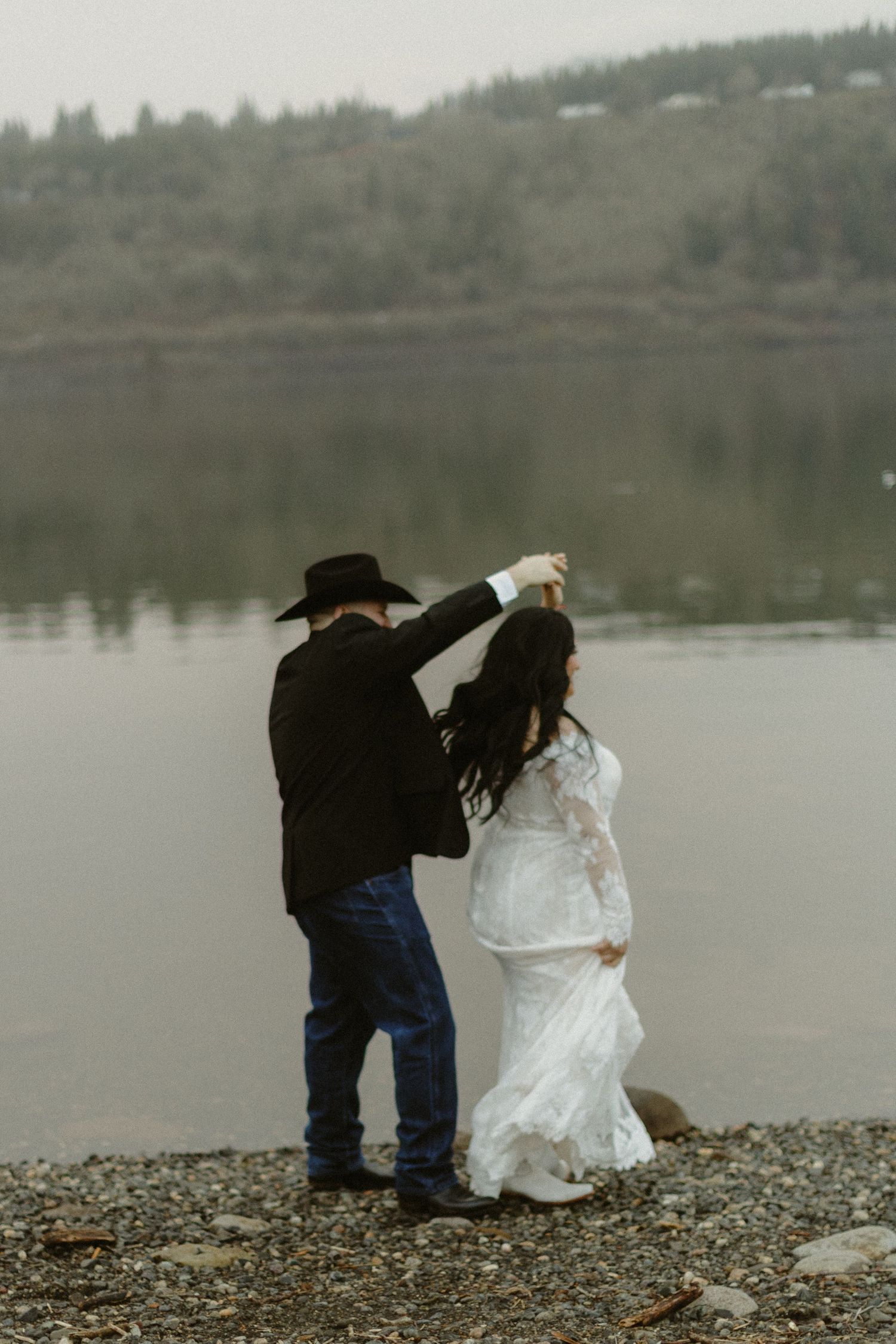 Groom spinning bride in white lace wedding gown by a calm misty lake with forested hillside backdrop.