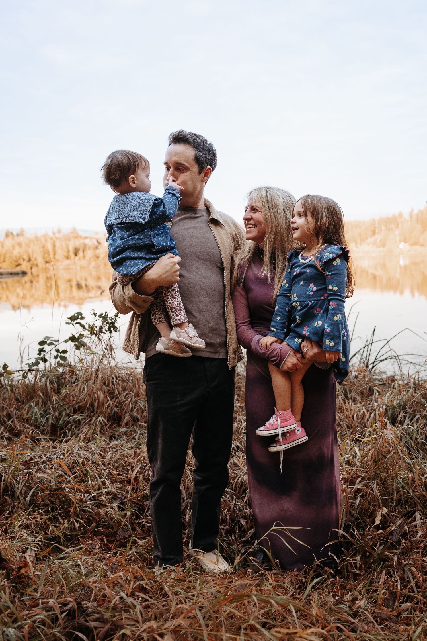 Family of four posing near autumn lake, parents holding two young daughters in blue floral dresses.