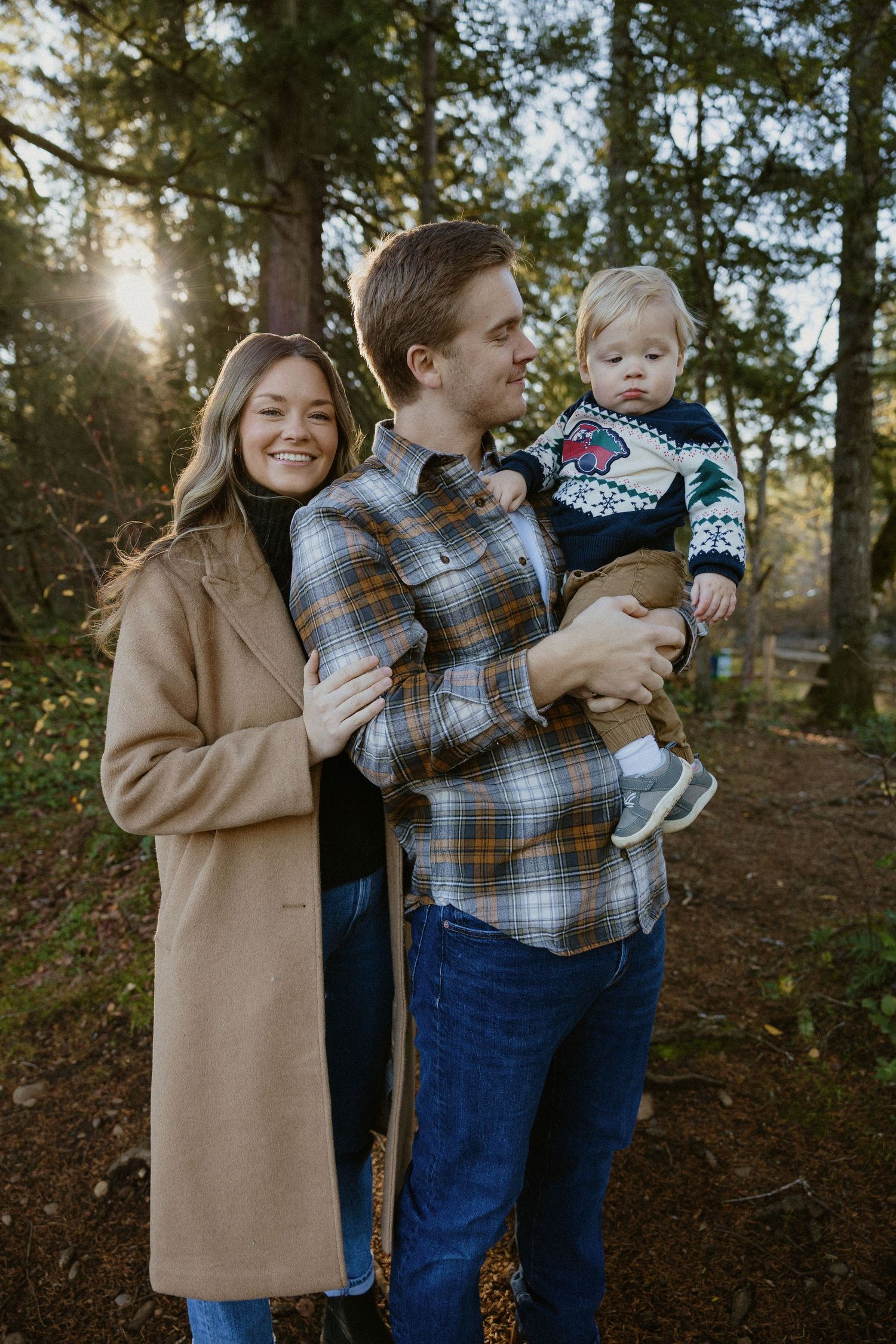 Smiling couple holding toddler in Christmas sweater during golden hour family portrait in woods.