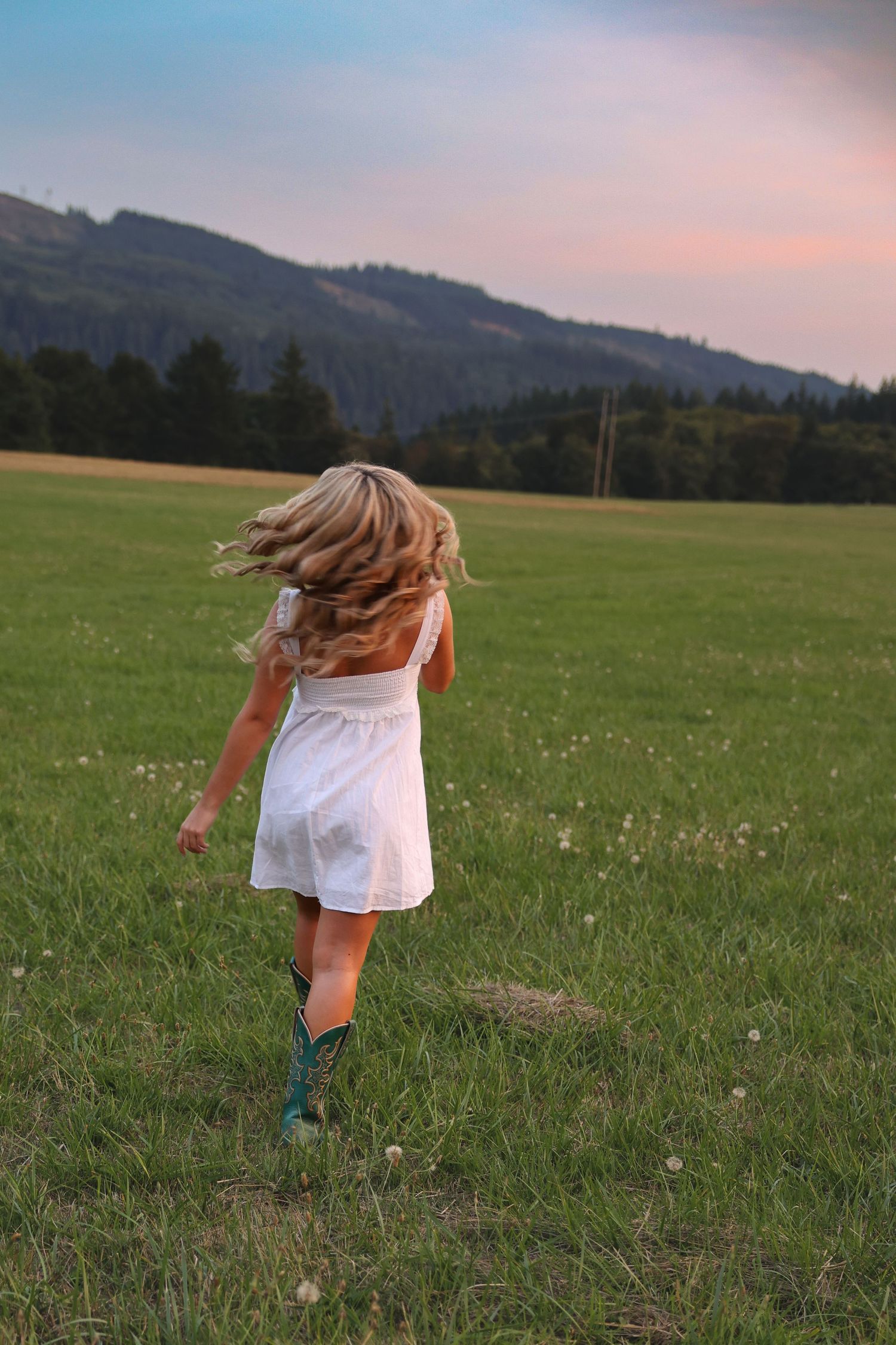 Young girl in white dress and boots running through green meadow with mountain backdrop at sunset.
