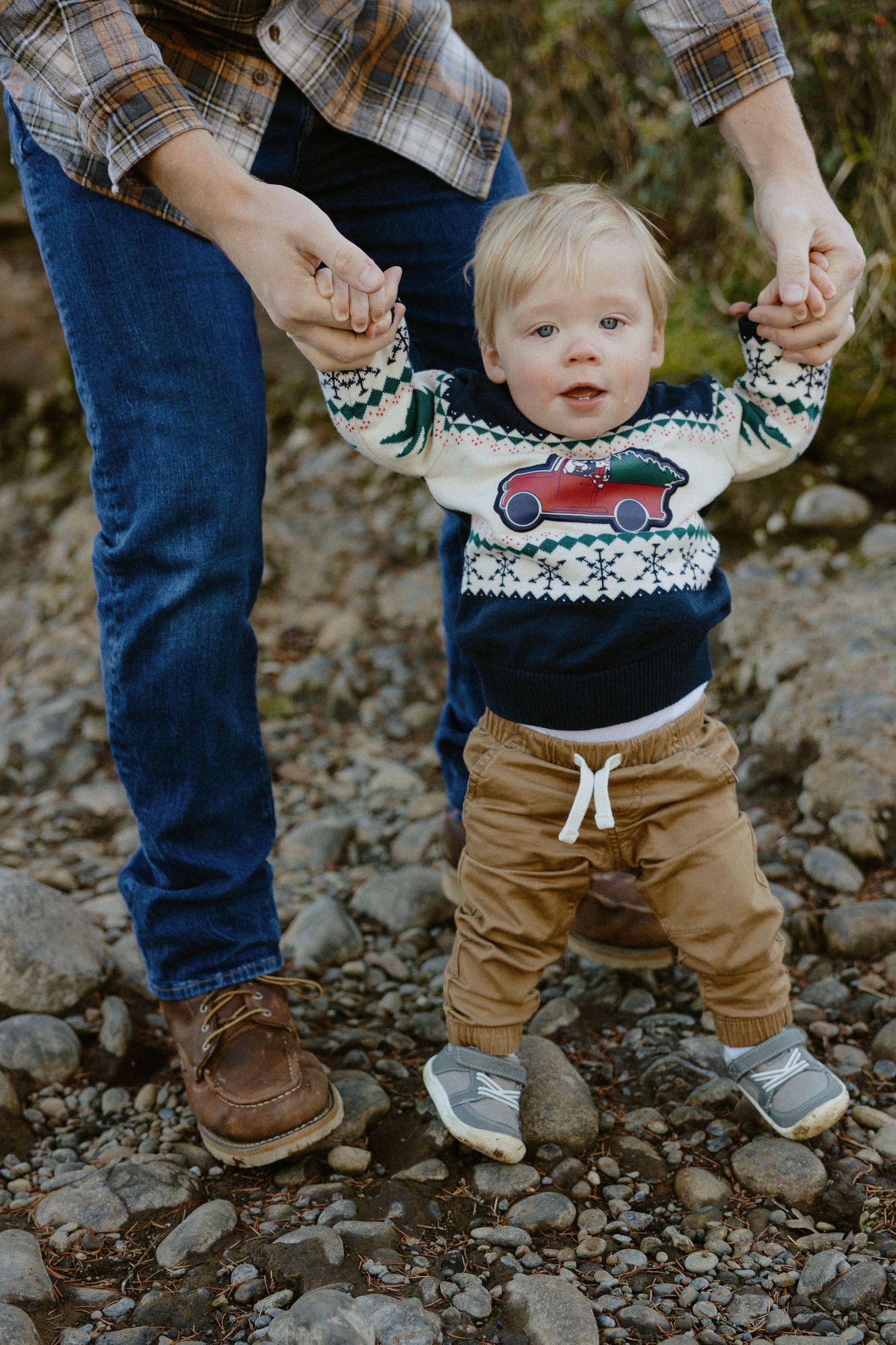 Adorable blonde toddler in a Christmas sweater learning to walk with parent's support outdoors.