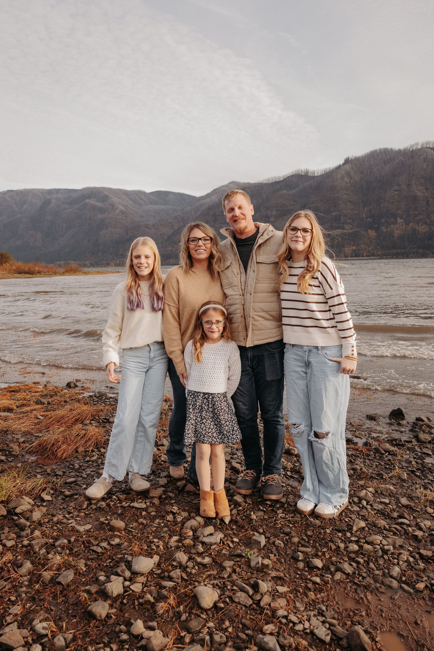 Family of five poses on a rocky shoreline with mountains and river in the background.