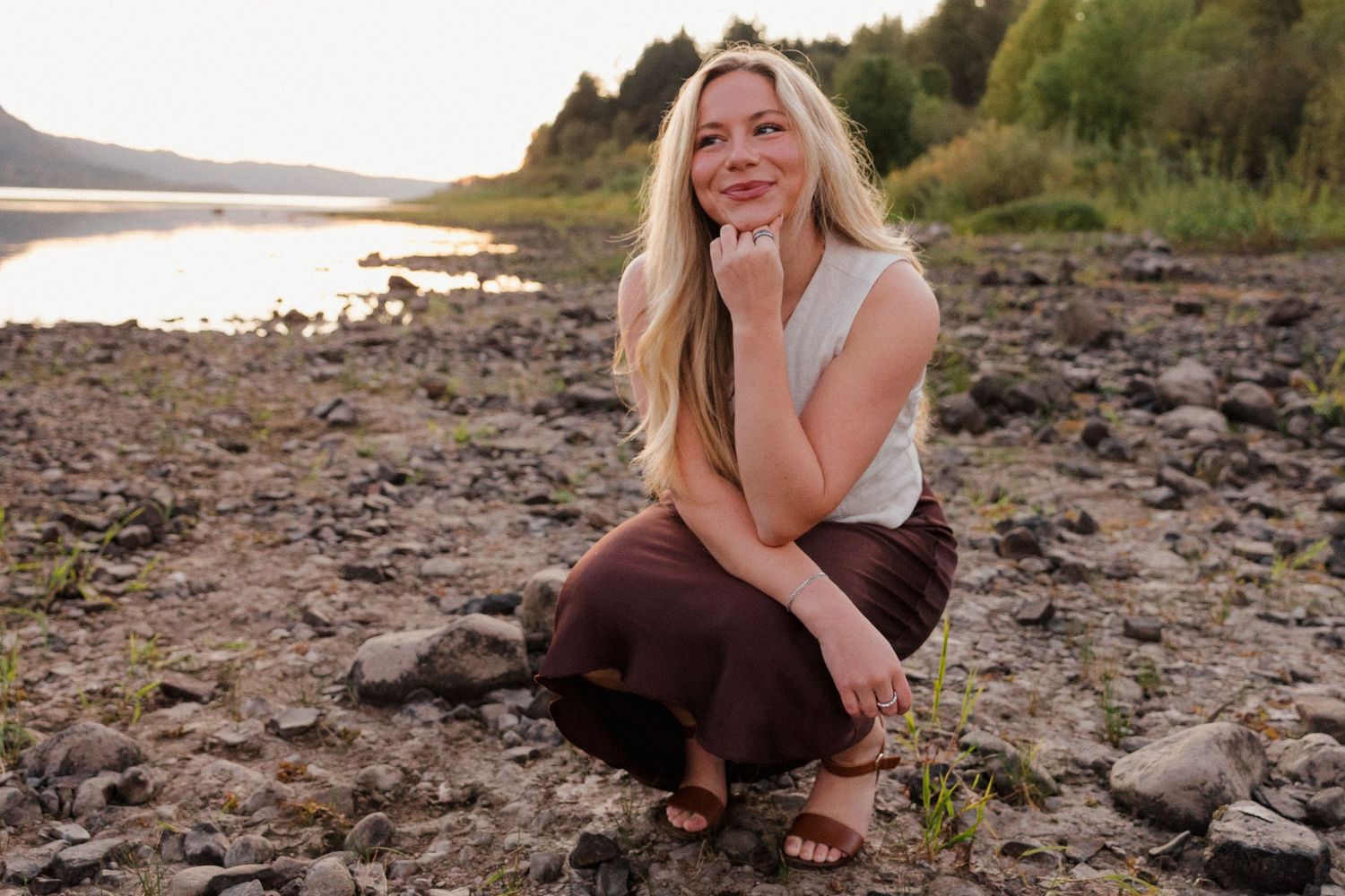 Smiling blonde woman crouching on rocky shore by a peaceful mountain lake at golden hour.