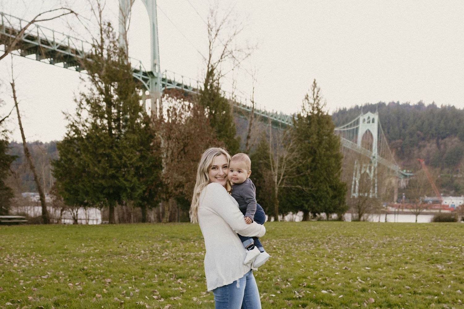 Blonde mother in white sweater holding infant with St. Johns Bridge visible in background on cloudy day.