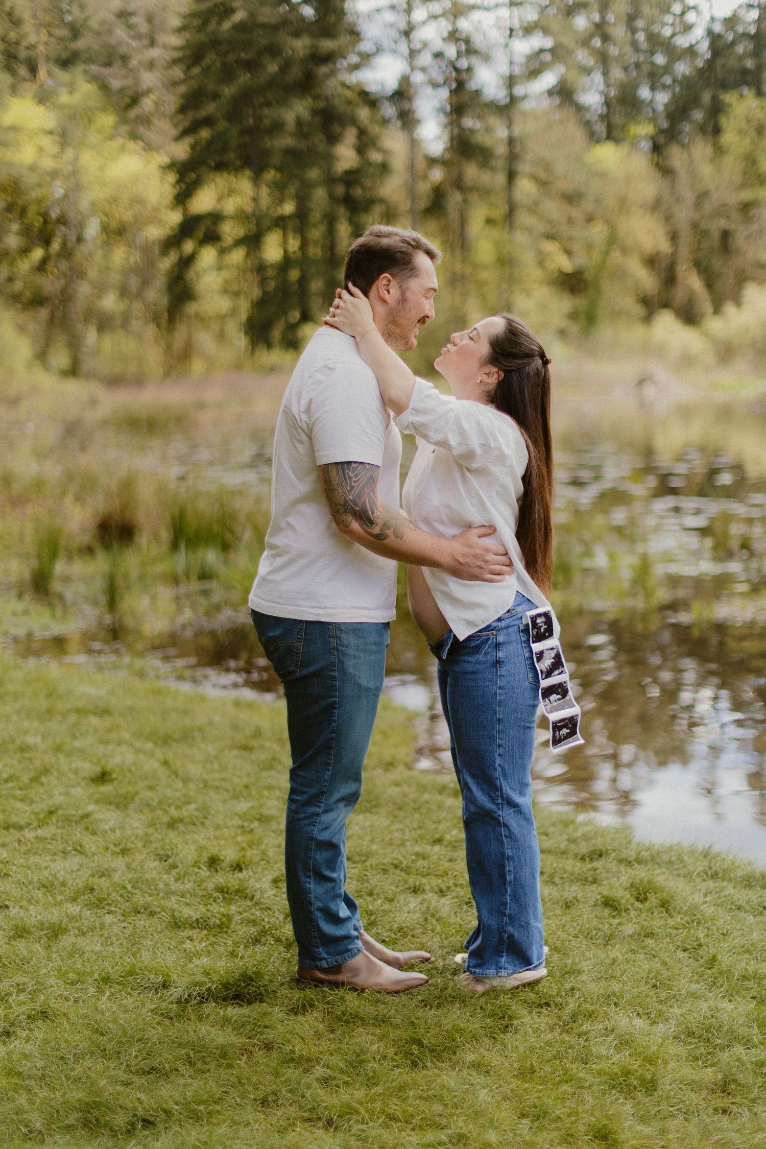 Couple sharing a joyful moment holding ultrasound photos by a serene pond surrounded by lush greenery.