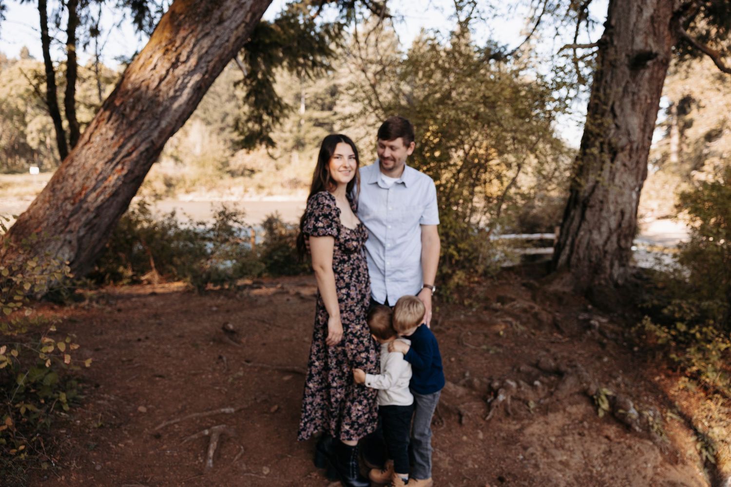 Family of four posing outdoors among tall trees on red dirt path in casual attire.