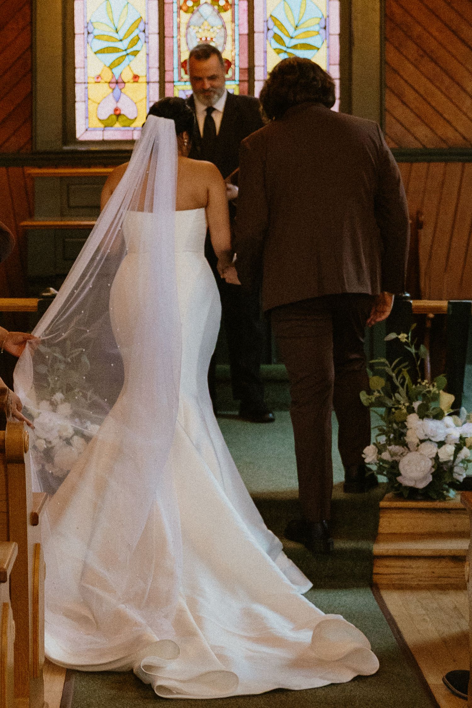 Bride in white mermaid gown with veil walks down church aisle toward officiant during wedding ceremony.