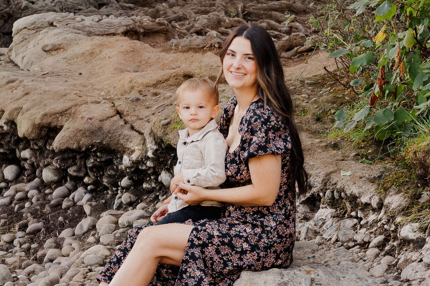 Brunette mother in floral dress smiling with toddler on rocky outdoor setting.