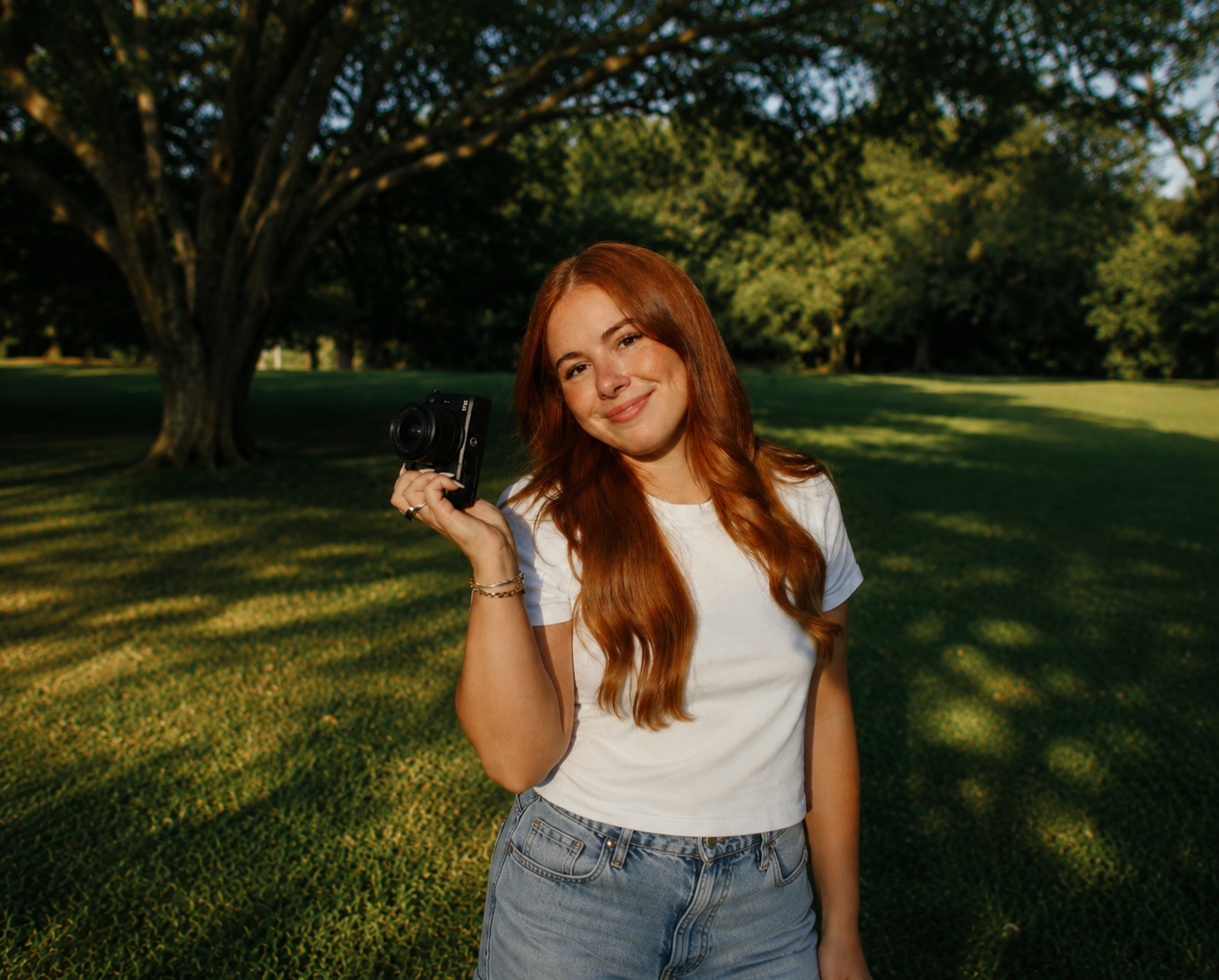 Young woman with red hair holding a camera, smiling outdoors in a sunny green park setting.