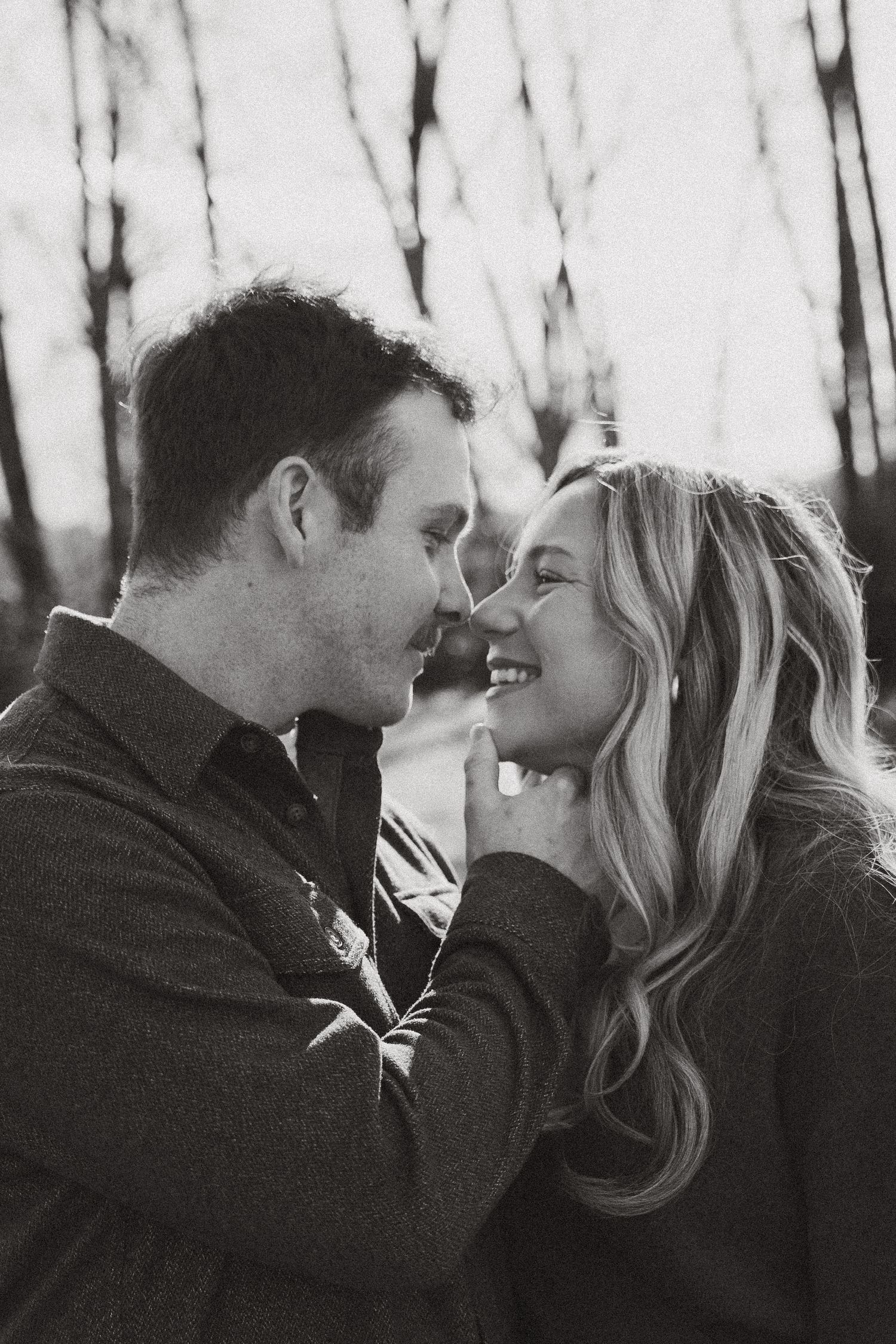 Black and white photo of couple smiling and almost kissing during romantic outdoor engagement shoot.