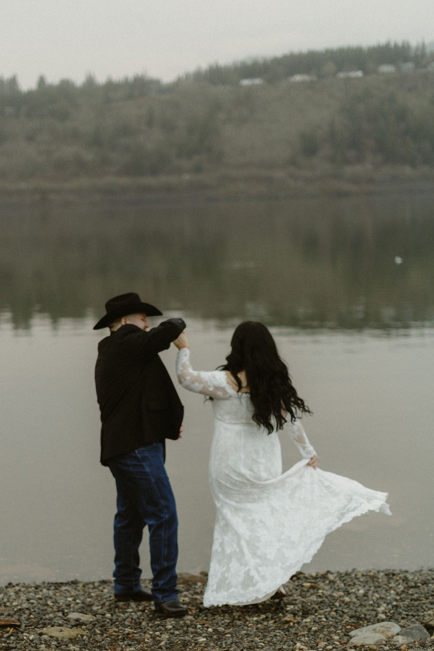 Groom in black cowboy hat twirling bride in flowing white lace gown on rocky lakeside shoreline.