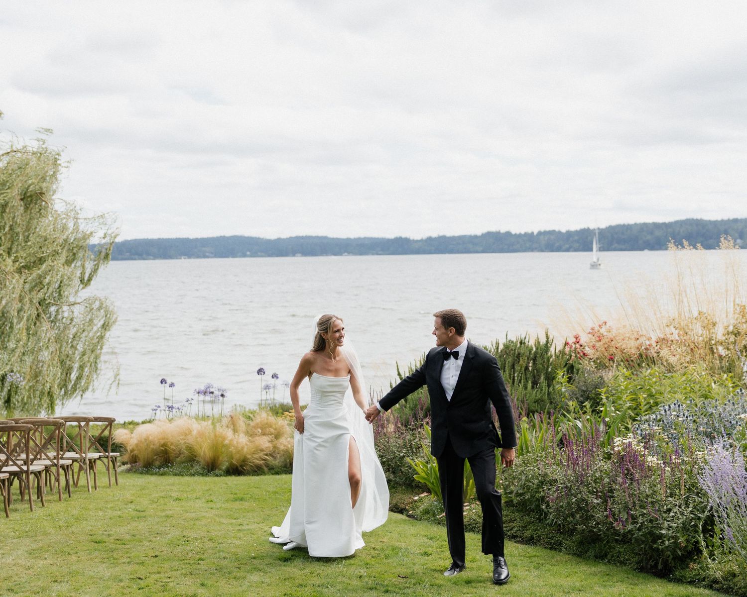 Bride and groom holding hands walking by a lakeside garden at an outdoor wedding ceremony.