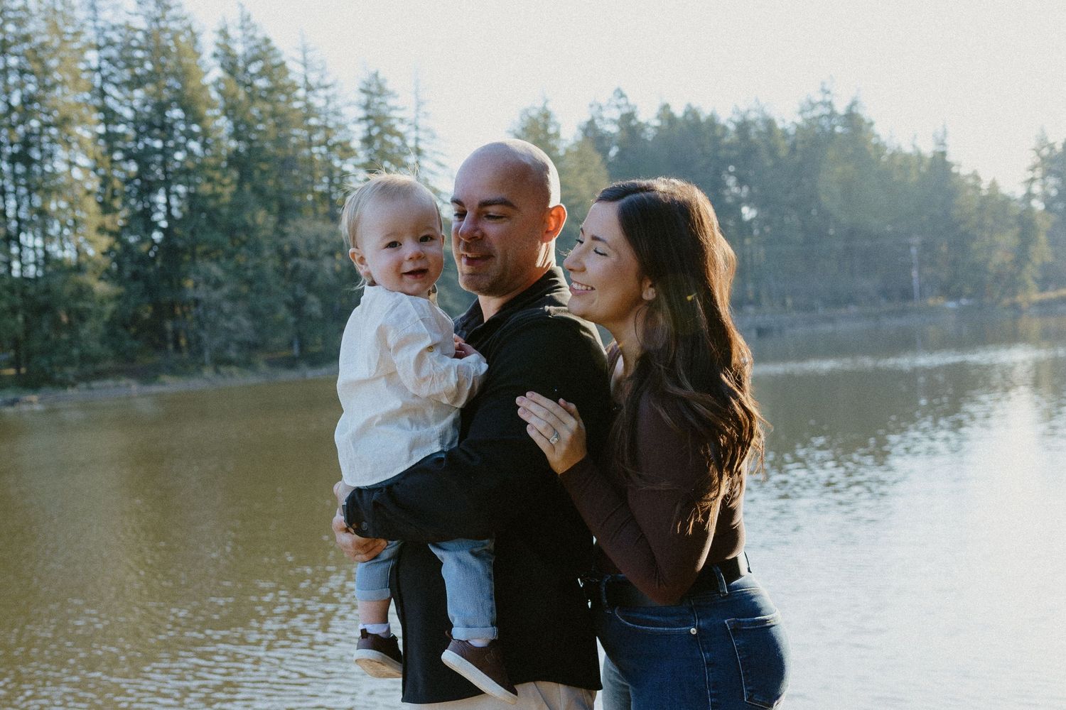 Parents smiling and holding their laughing baby outdoors by a sunlit lake with evergreen trees.