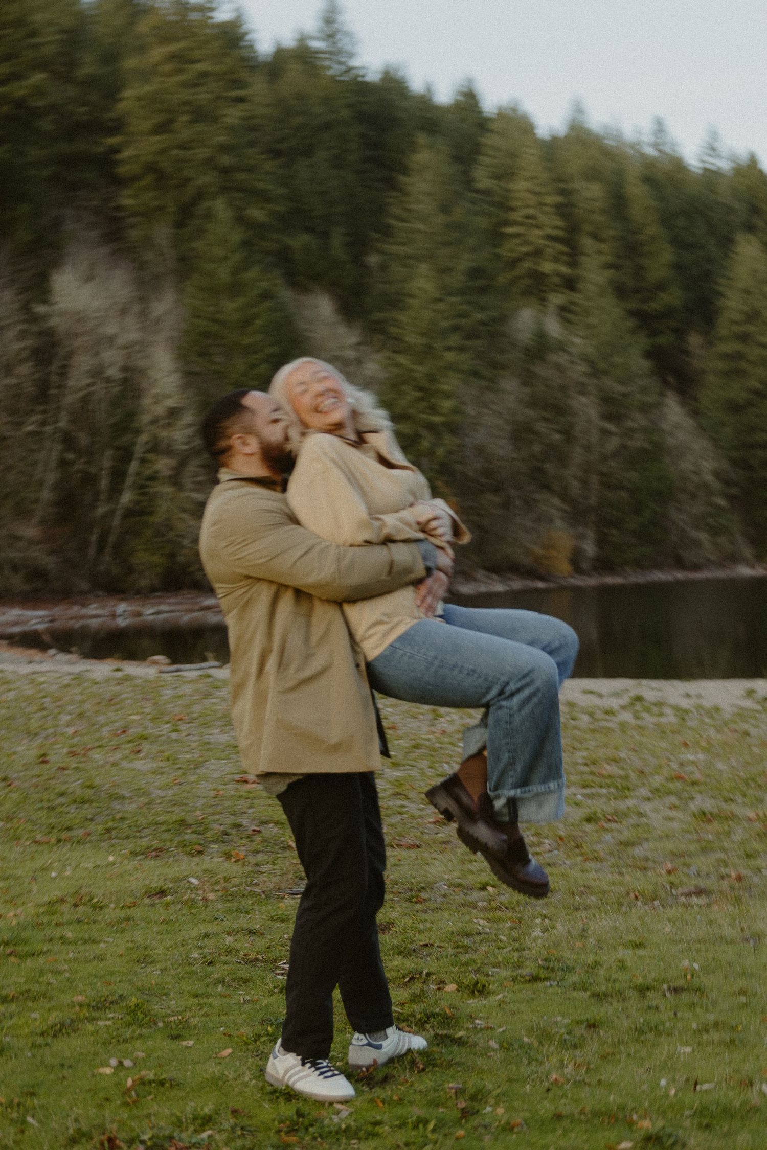 Man lifting woman in his arms outdoors near a lake with evergreen trees in background.