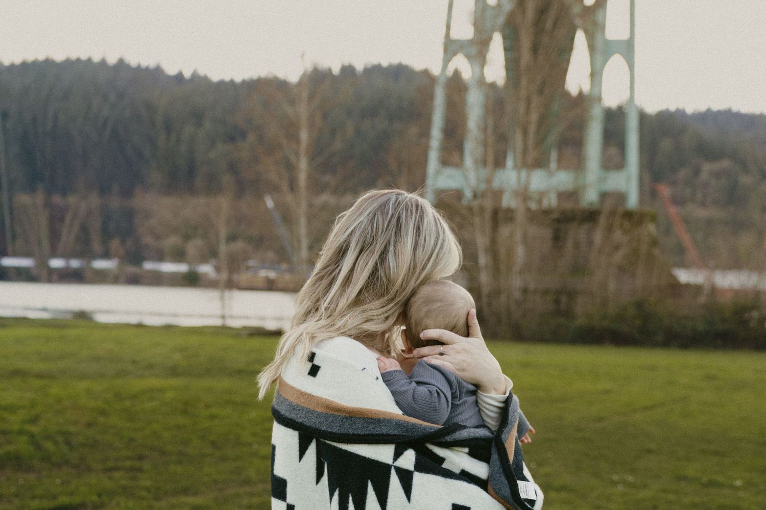 Mother in geometric patterned blanket cradling newborn baby with St. Johns Bridge visible in background.