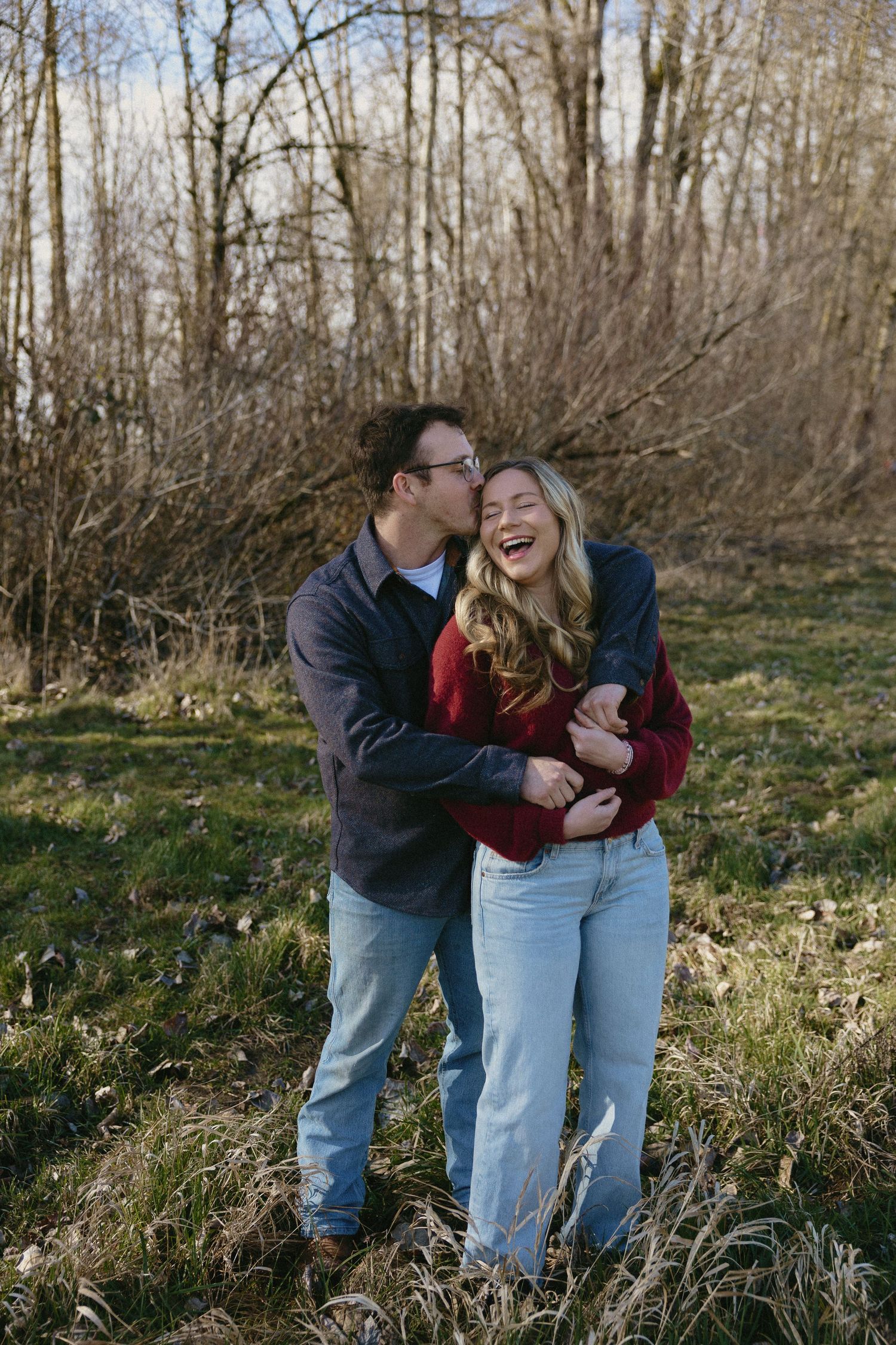 Couple laughing and embracing outdoors in a wooded field during winter engagement photo session.