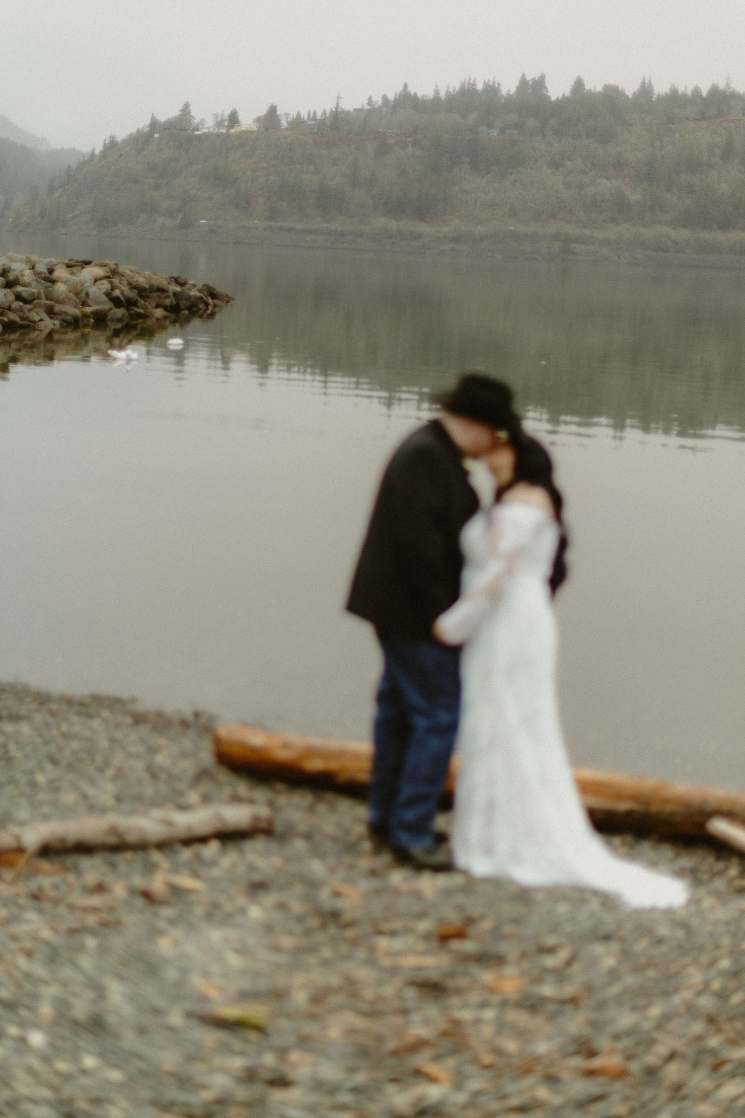 Bride and groom sharing a romantic embrace by a misty lake with forested hills in the background.