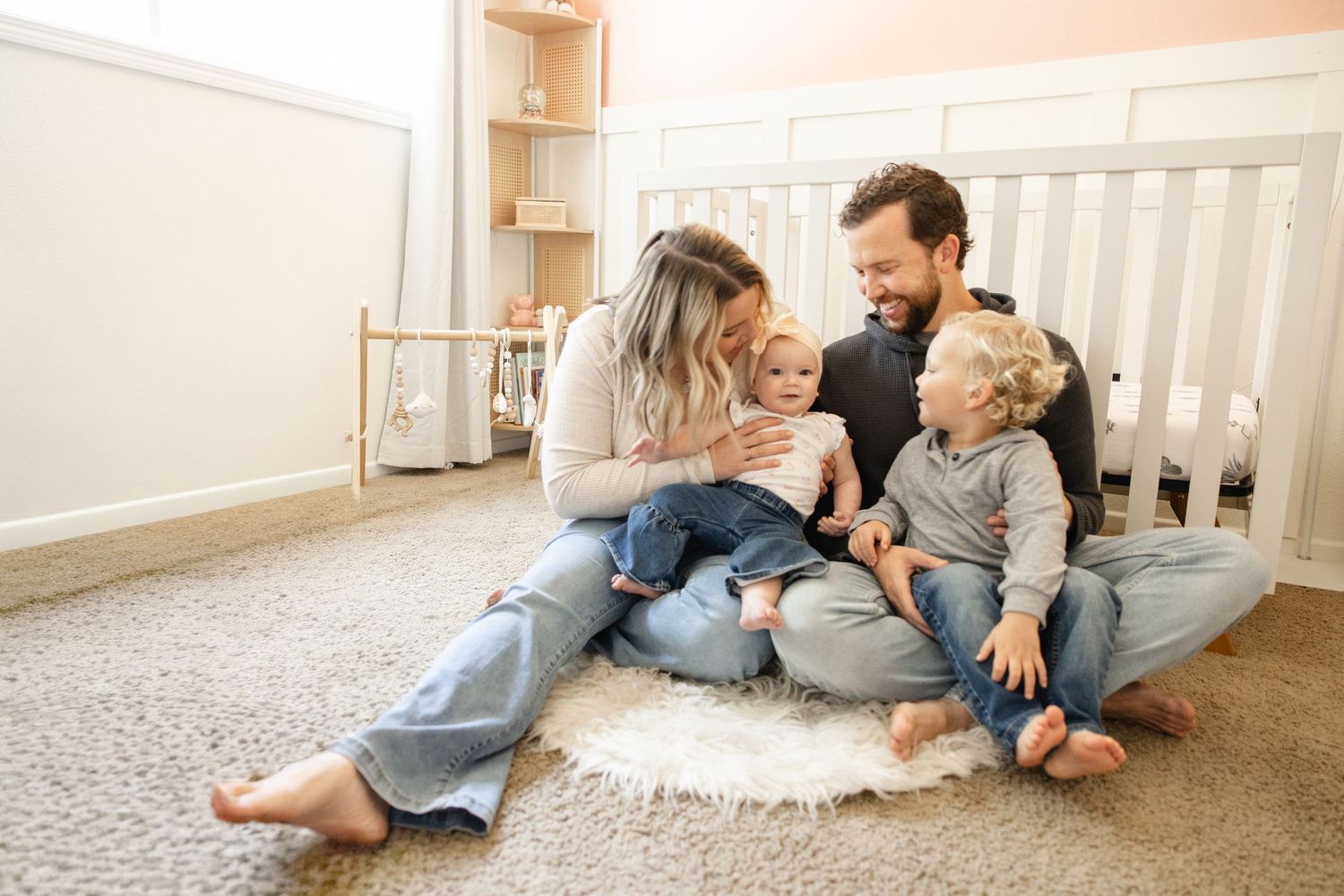 Happy family laughing together on nursery floor with two young children near white crib.