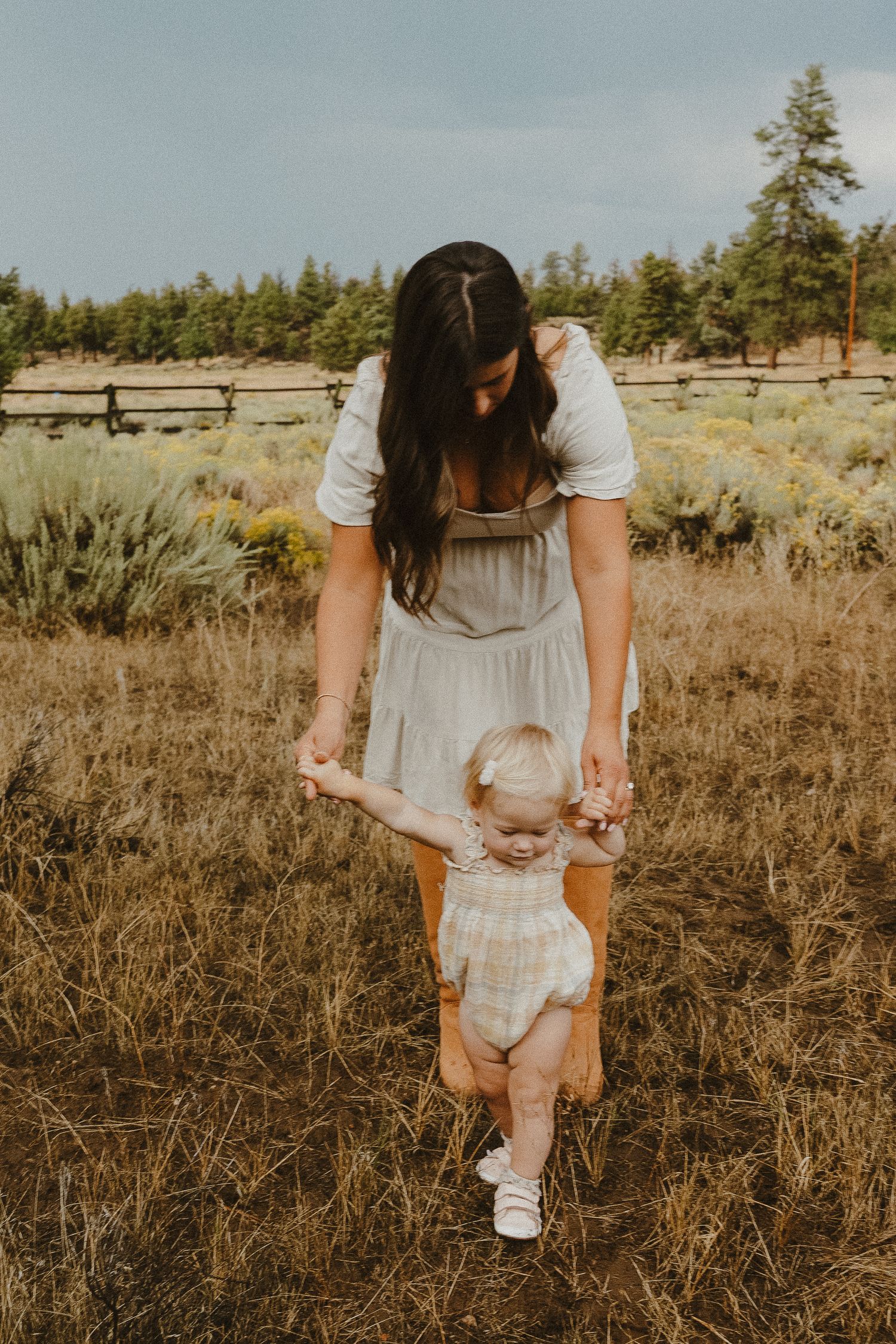 Mother in white dress helps baby take first steps in a rustic outdoor field with pine trees.