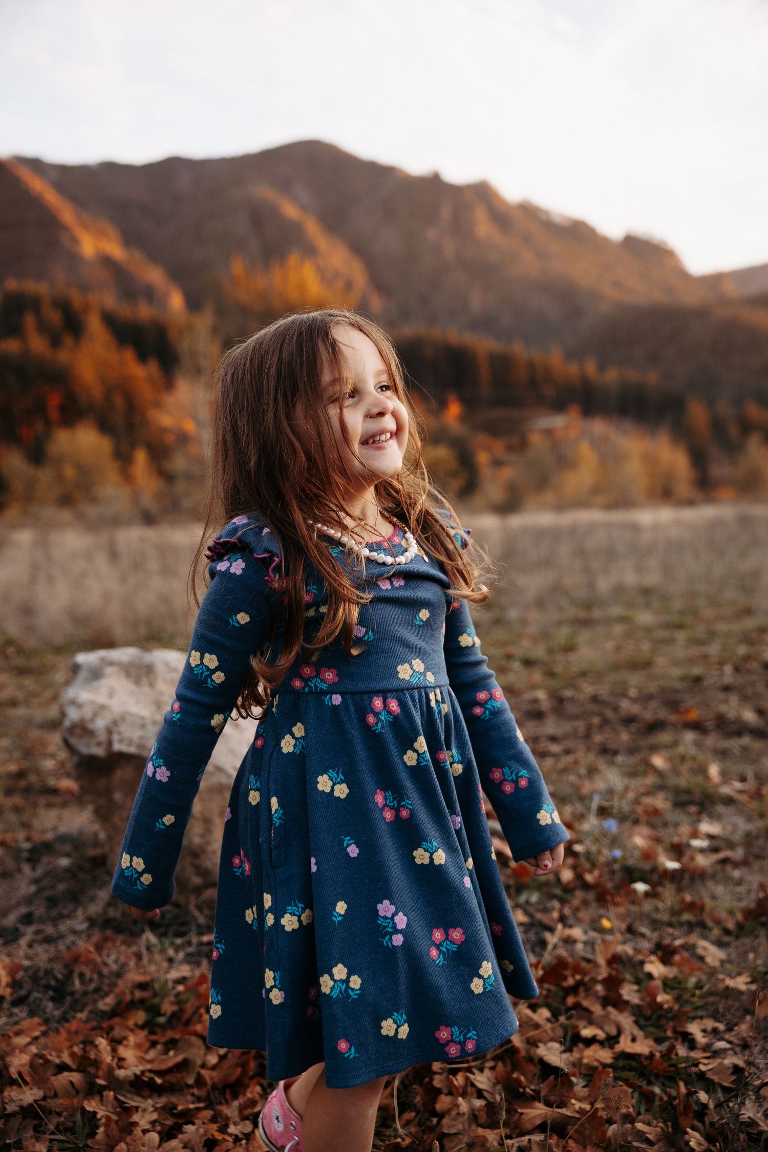 Joyful young girl in navy floral dress spinning in autumn leaves with mountain forest backdrop.