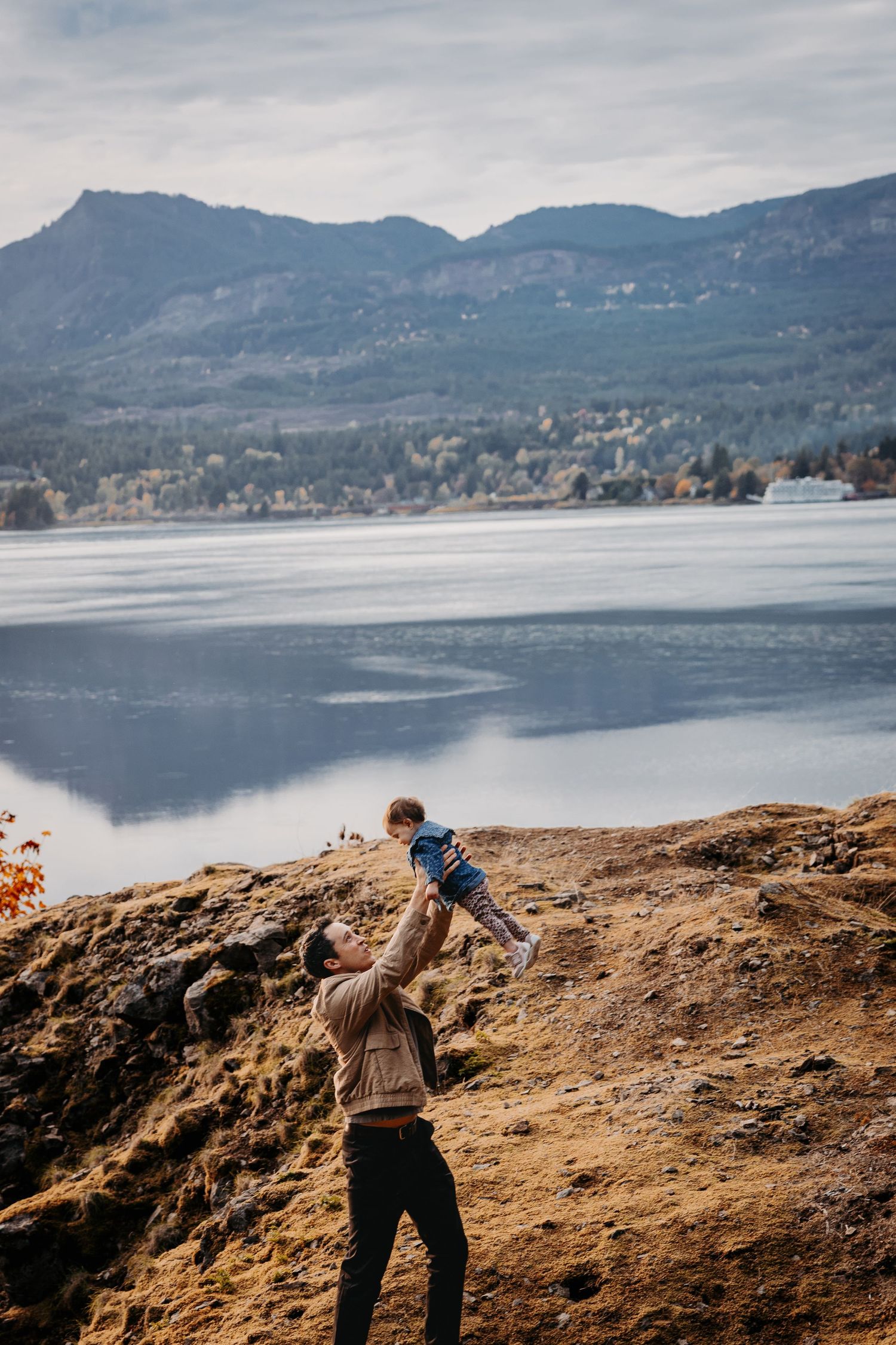Father lifting baby in the air with scenic mountain lake and cloudy sky in background.