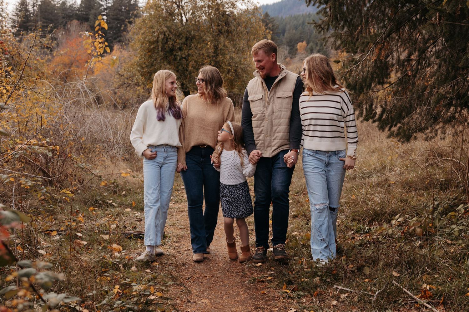 Family of five walking on autumn trail path through golden foliage in casual fall outfits.