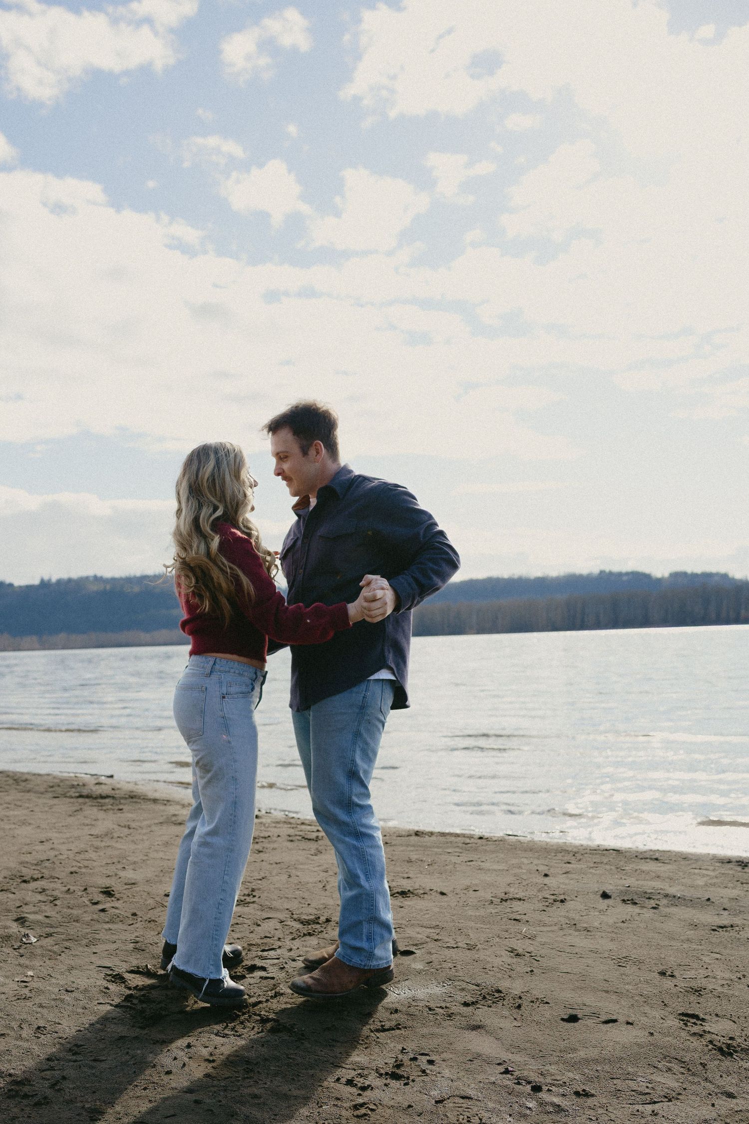 Couple dancing together on a sandy beach by a lake under a partly cloudy sky in fall clothing.