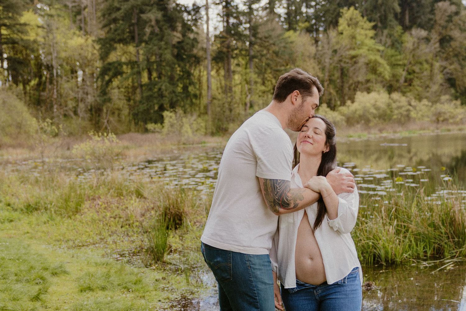 Man tenderly embracing pregnant woman from behind near a tranquil woodland pond.